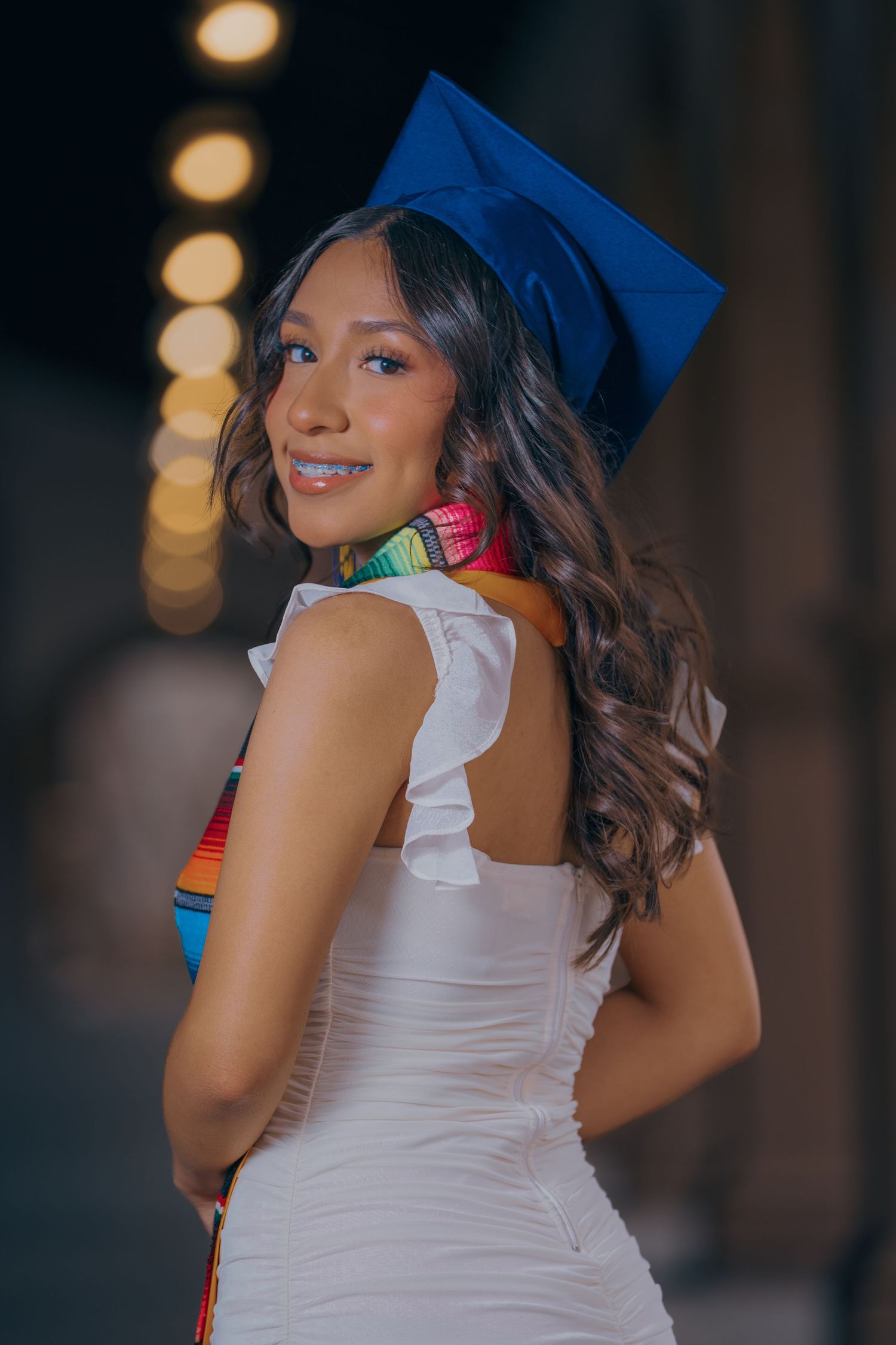 Graduate in white dress and blue cap poses for graduation photos with bokeh lights in background.