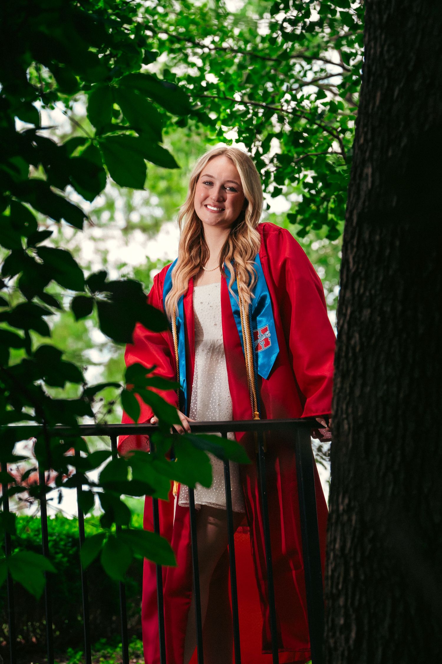 Graduate in bright red academic regalia poses among green foliage during outdoor graduation photo session.