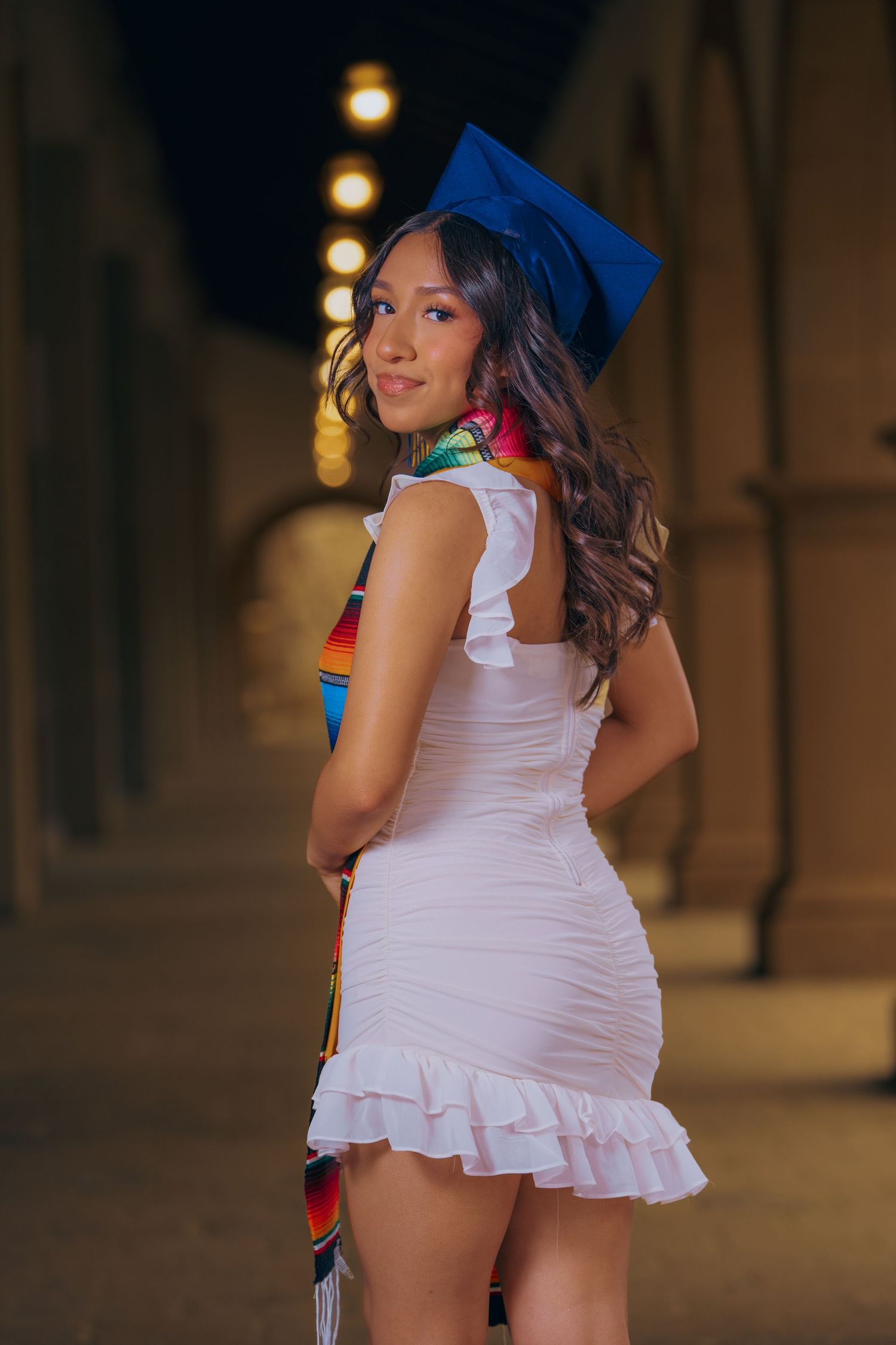 Graduate in blue cap and white dress poses in arched hallway with decorative sash at sunset.