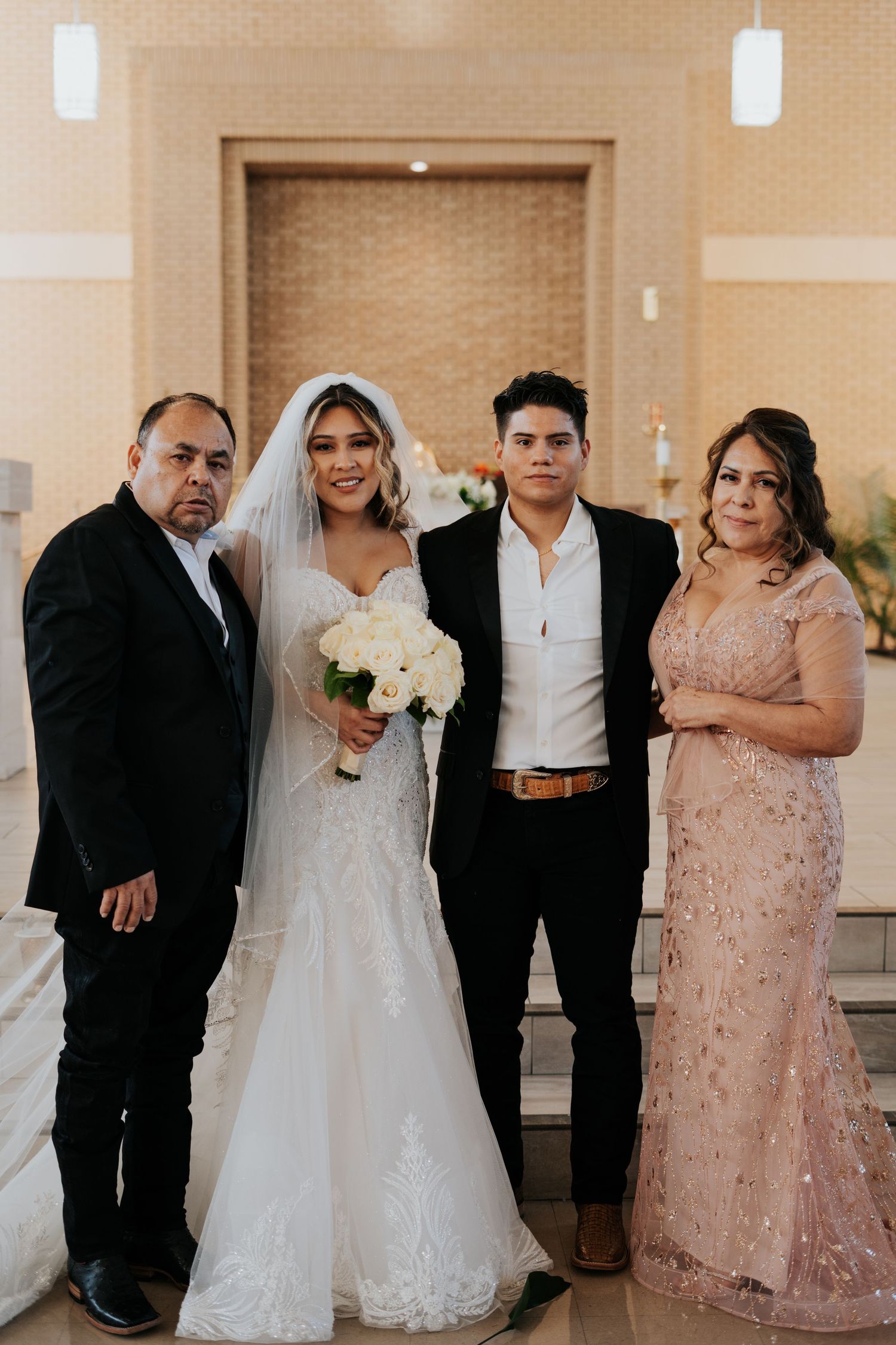 Wedding party poses together in formal attire against neutral background at indoor venue.