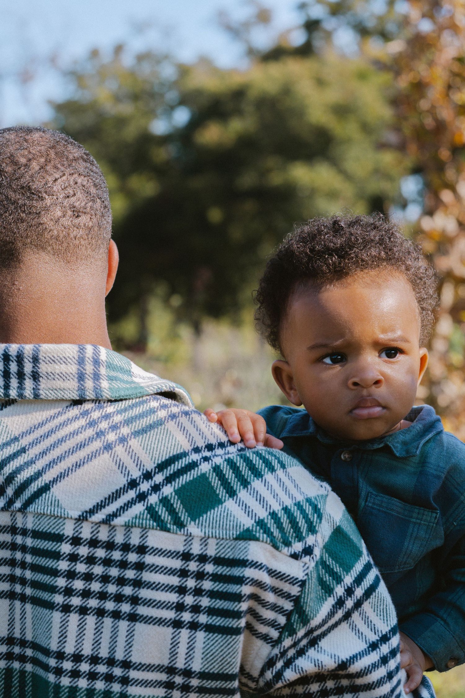 A man holds a curious baby outdoors, with the infant looking over his shoulder at the camera.