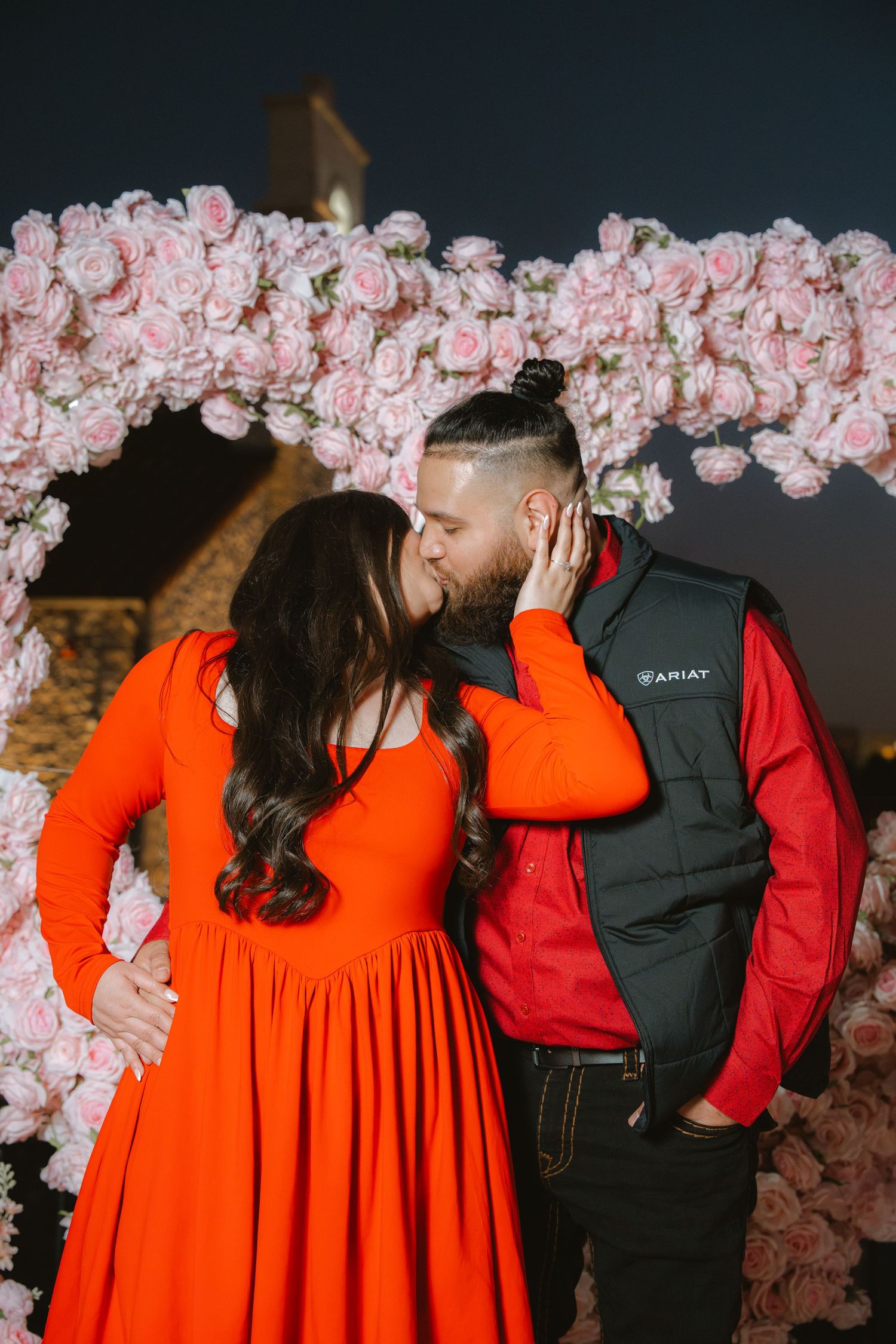 Couple sharing a kiss in front of a floral arch of pink roses during an outdoor marriage proposal at night.