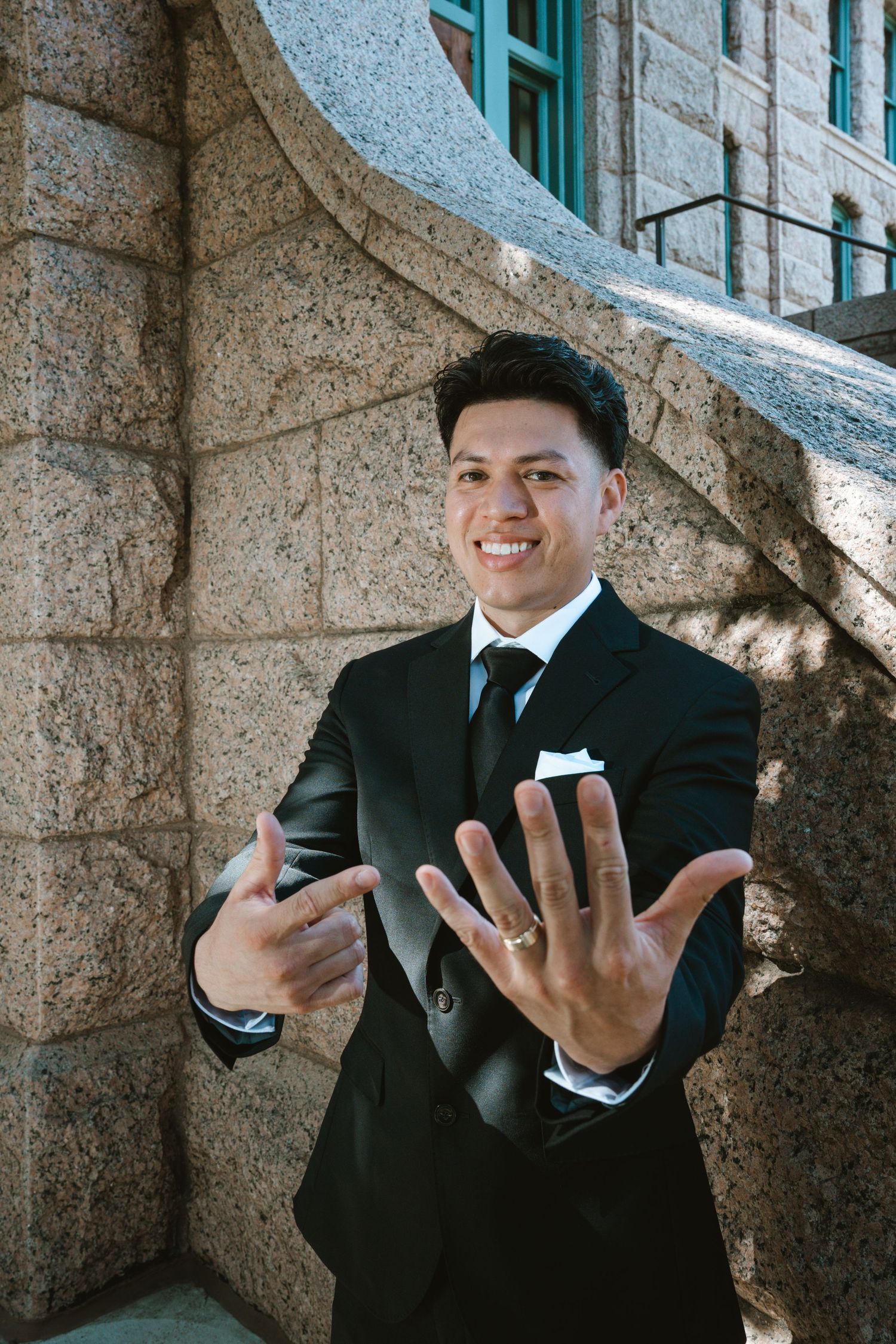 Smiling man in black suit displaying wedding ring outdoors near stone building.