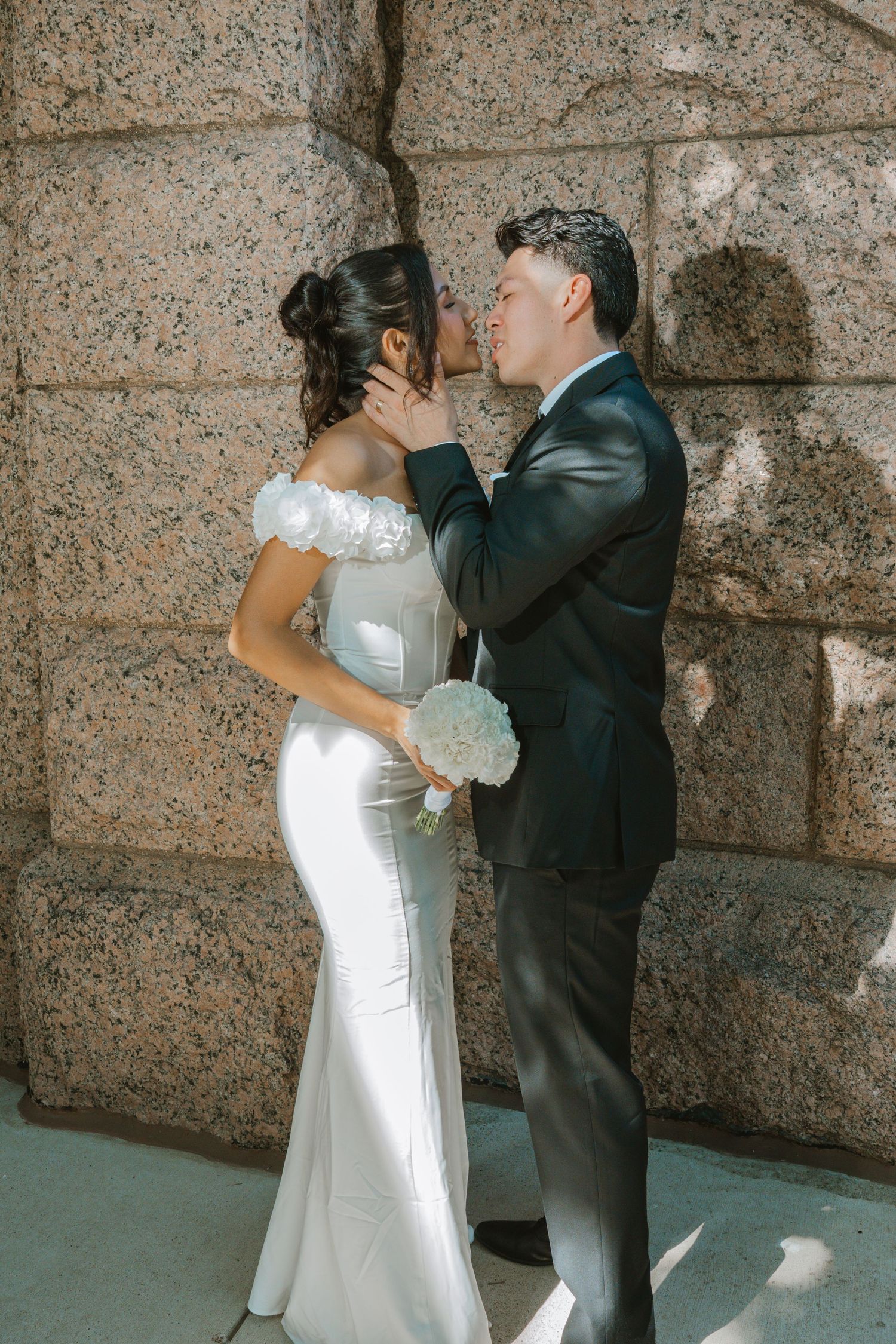 A bride and groom share a romantic kiss against a stone wall, she in a white satin gown holding a bouquet.