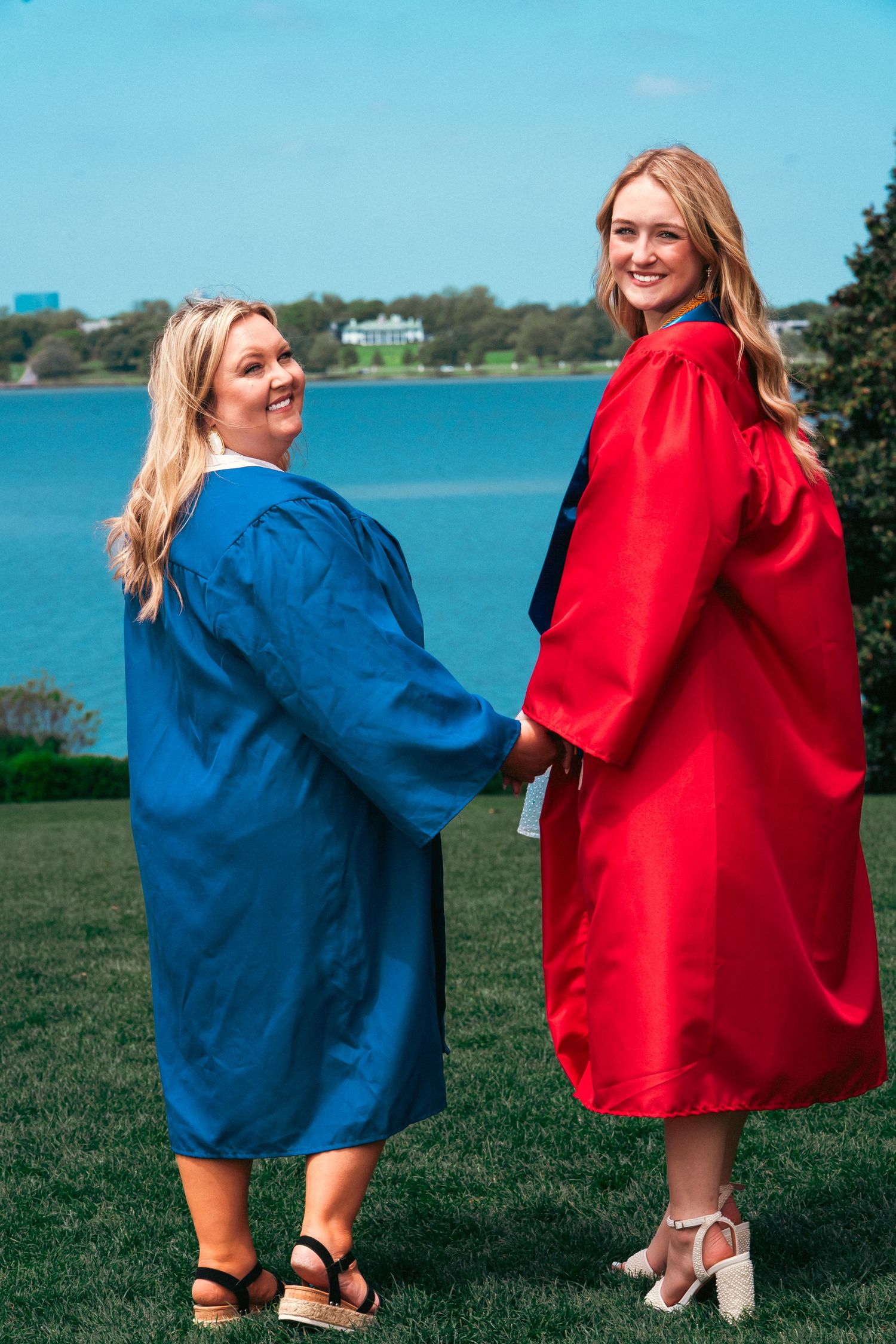 Two graduates in red and blue commencement robes pose together on grass overlooking a lake on a sunny day.