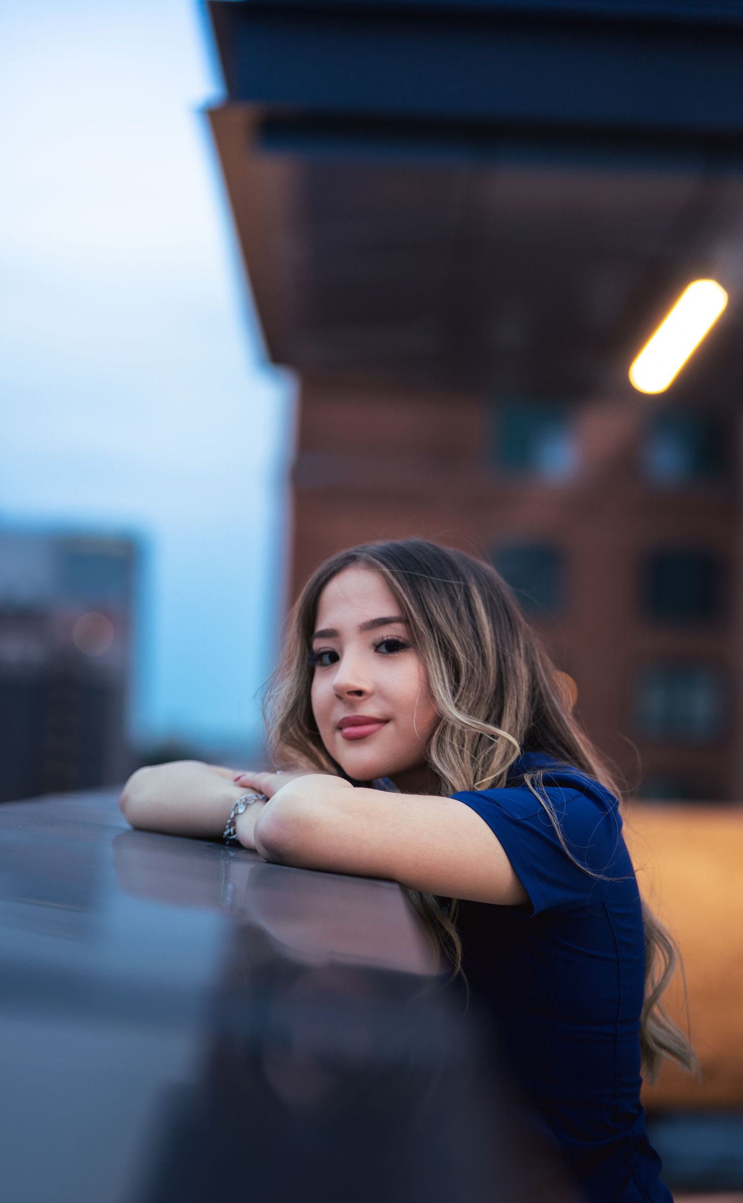 A person in a dark blue shirt leans on a surface during dusk with warm lighting in an urban setting.