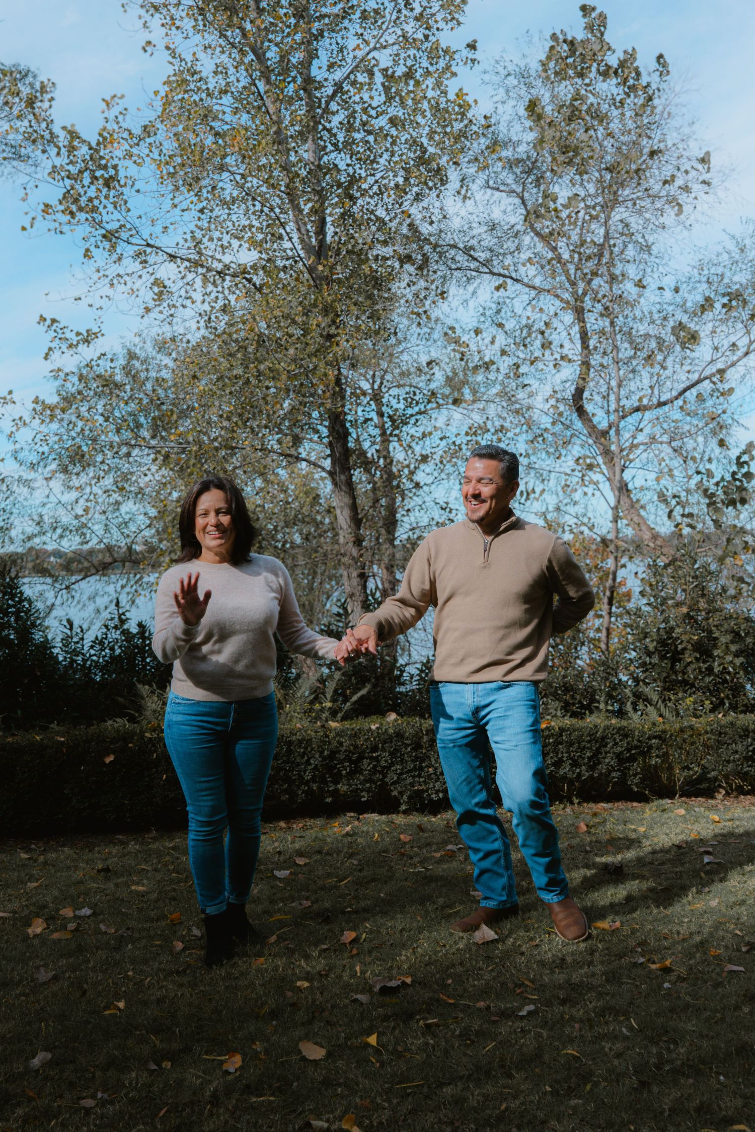 A couple playfully walks and dances on a grassy hillside with autumn trees in the background.