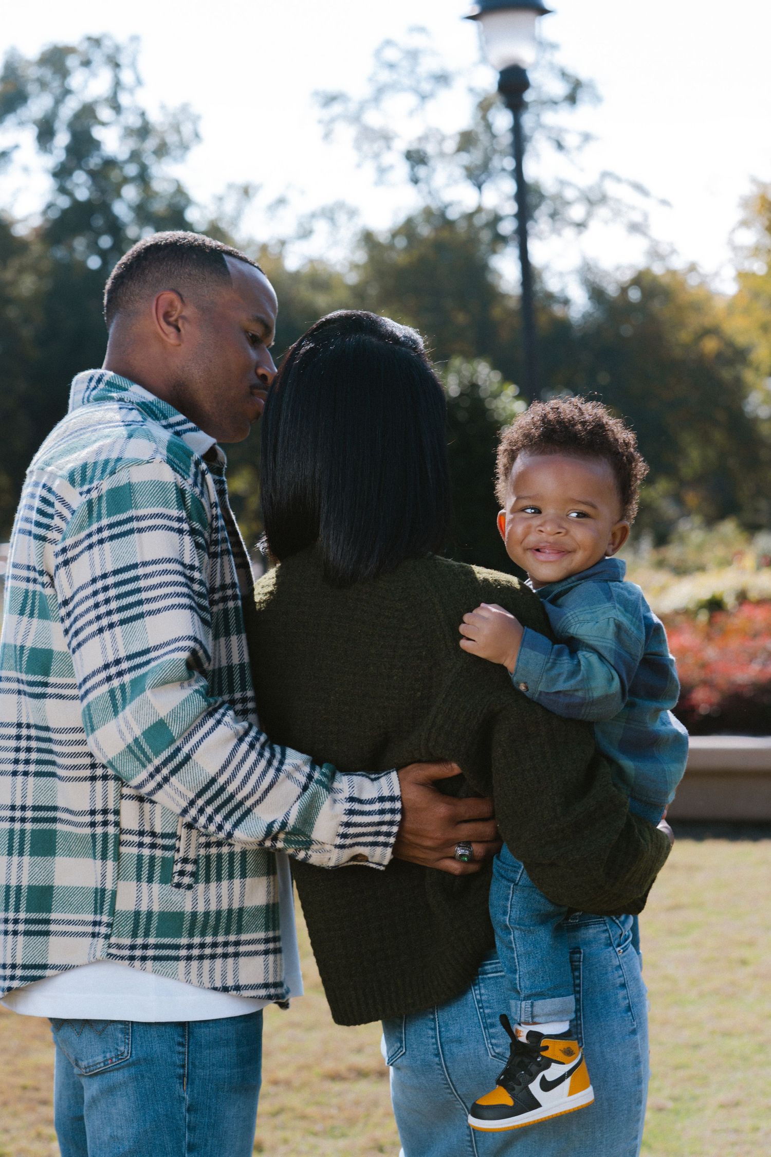 A loving Black couple shares a kiss while holding their smiling toddler in a sunny park setting.
