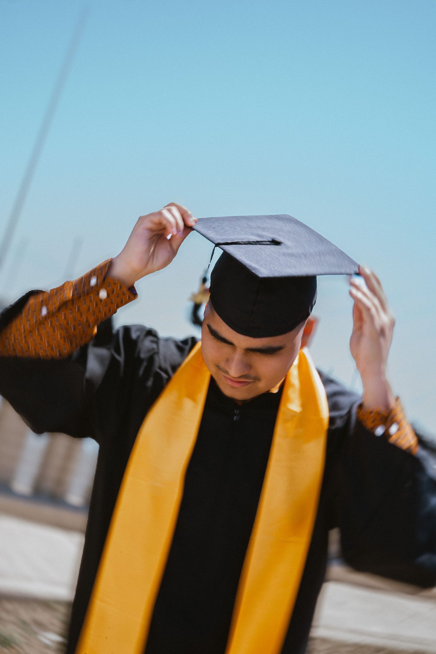 Young male graduate in black cap and gown with gold stole adjusting his mortarboard under blue sky.