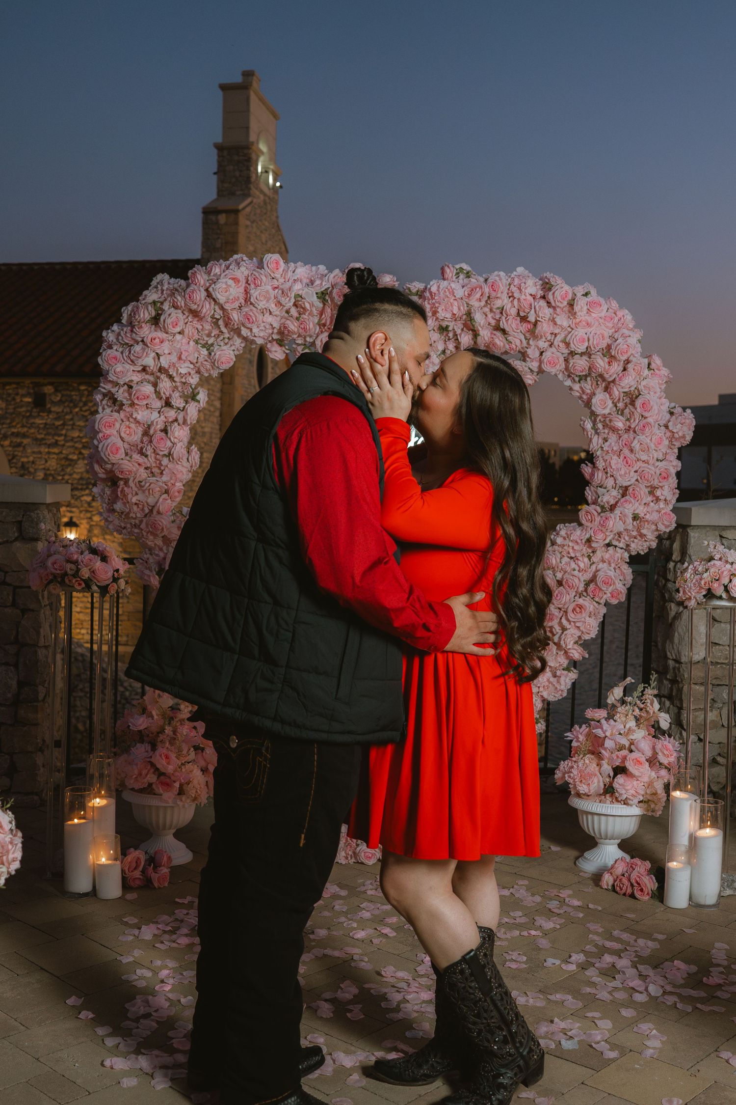 Couple kissing in front of a large pink flower heart arch with candles at dusk during a romantic proposal.