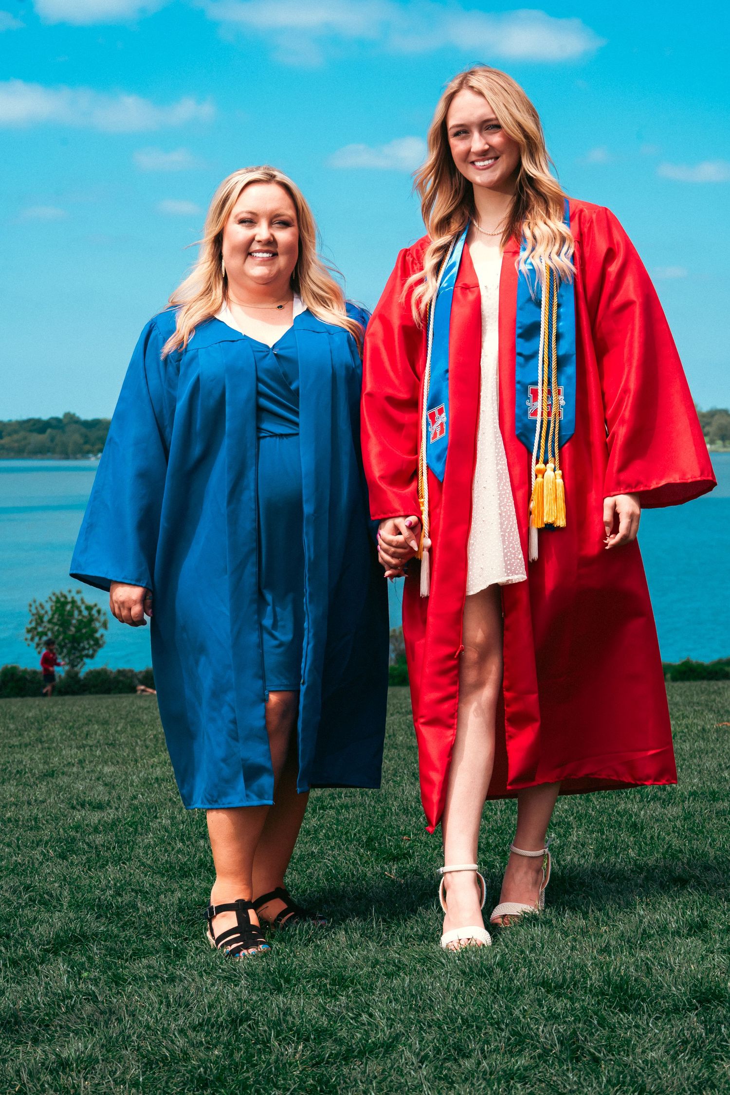 Two graduates in red and blue gowns pose on a grassy hill overlooking a scenic waterfront view.