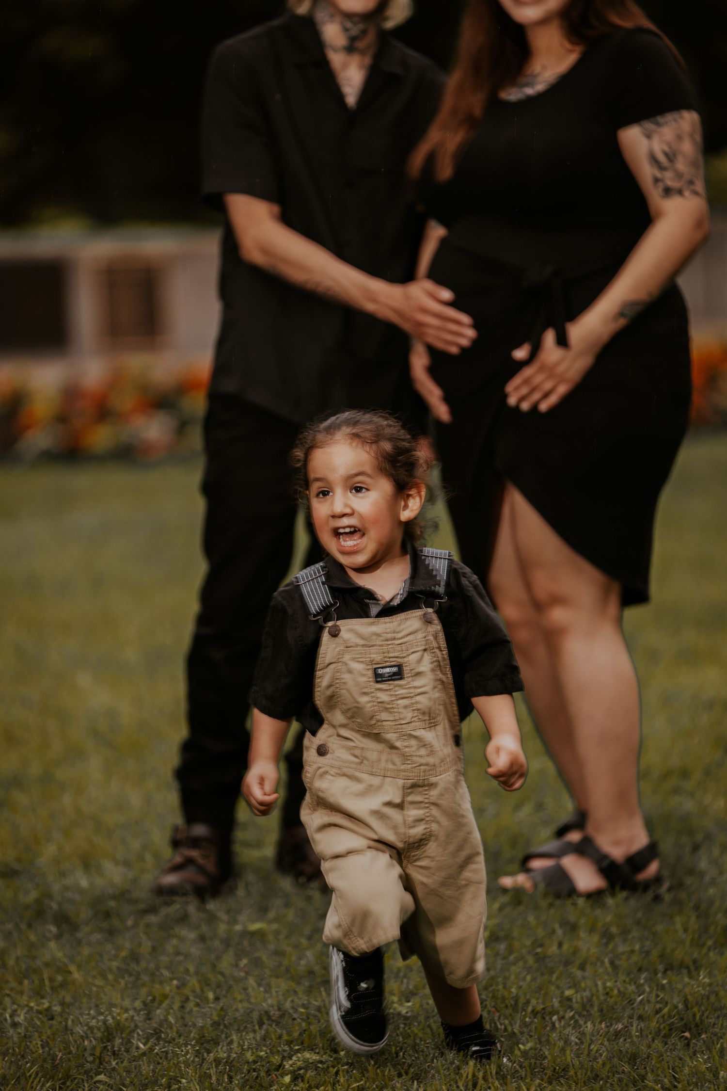 A small child in tan overalls runs playfully through grass while adults in black clothing watch nearby.