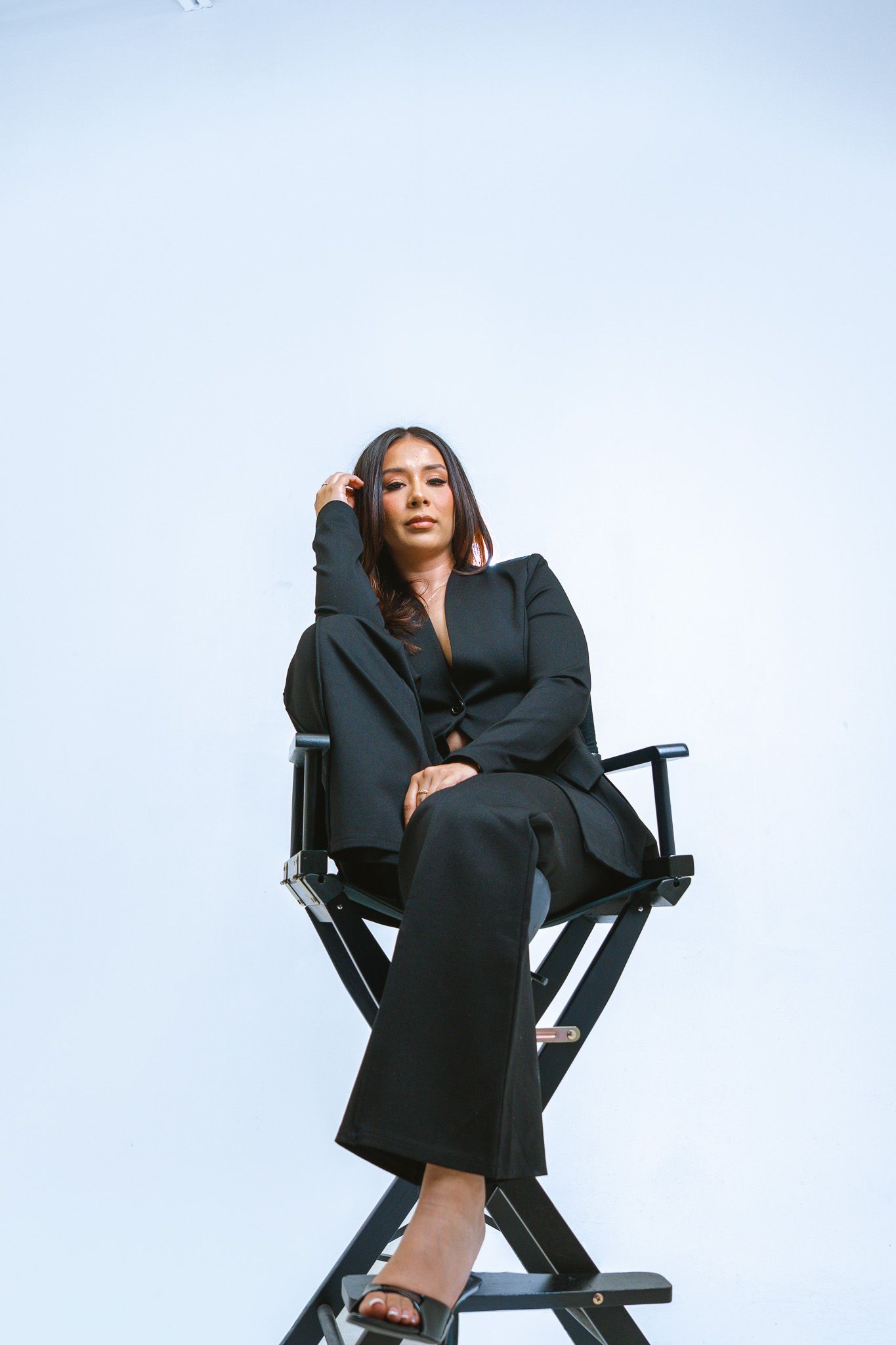 Professional fashion shoot of a model posing dramatically in a black outfit on a director's chair against white backdrop.