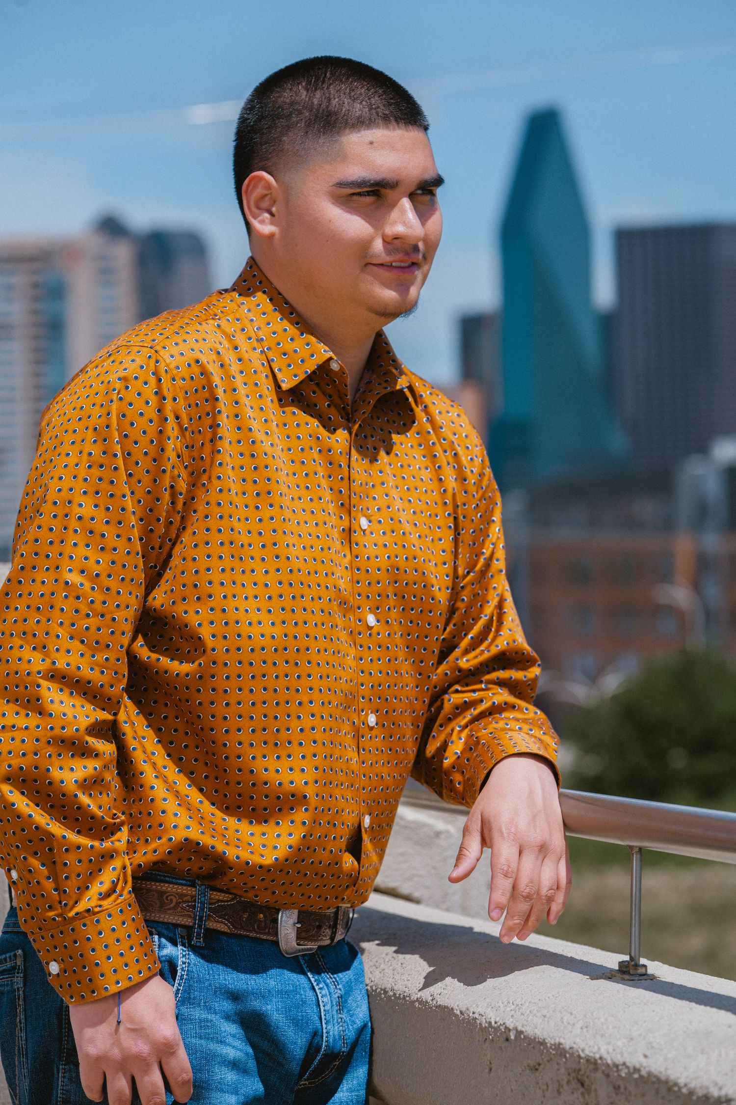 Young man in mustard yellow patterned shirt and jeans poses on urban rooftop with city skyline behind him.