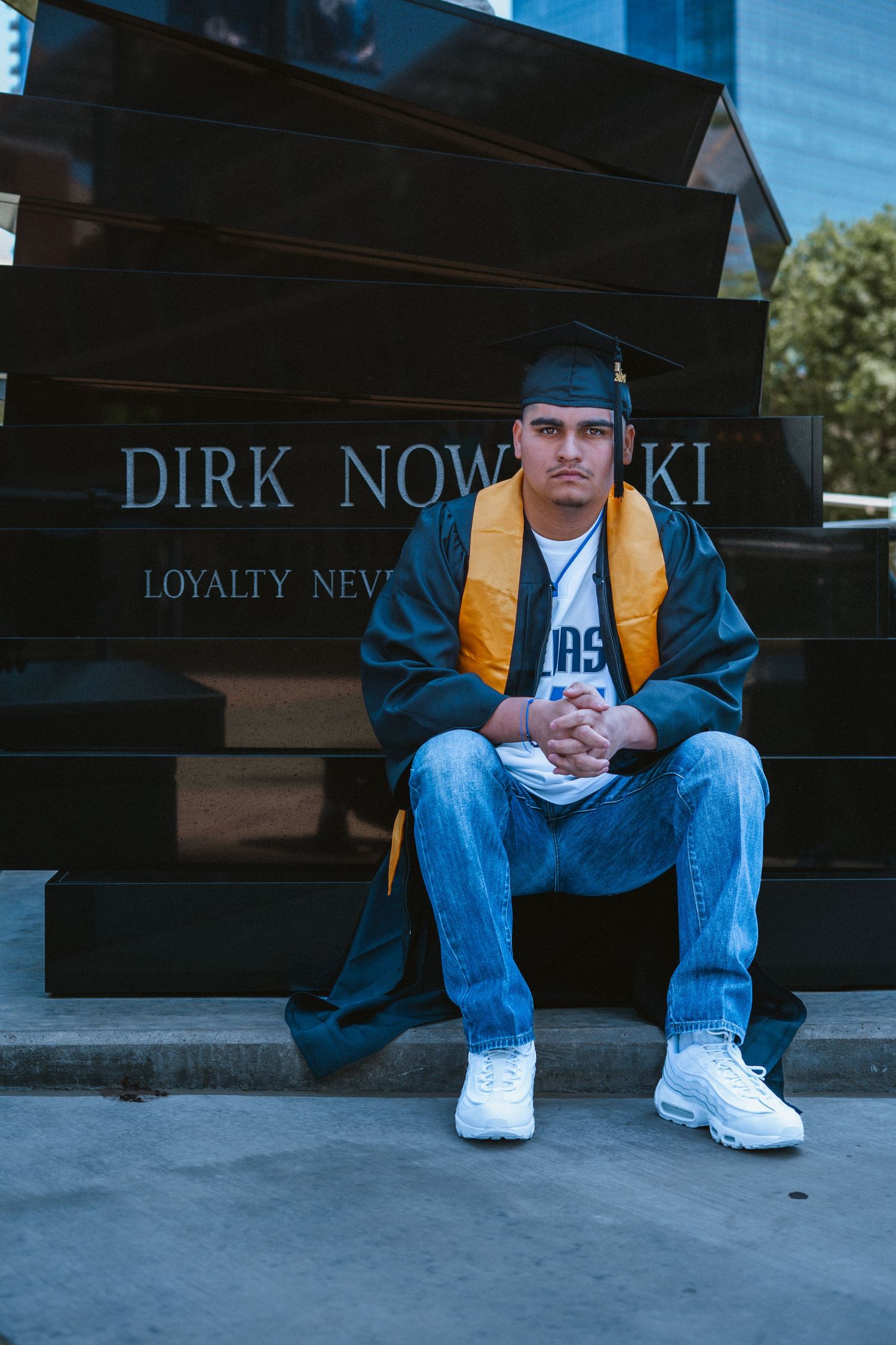 Graduate in cap and gown sits before Dirk Nowitzki monument in Dallas, wearing gold stole and white sneakers.