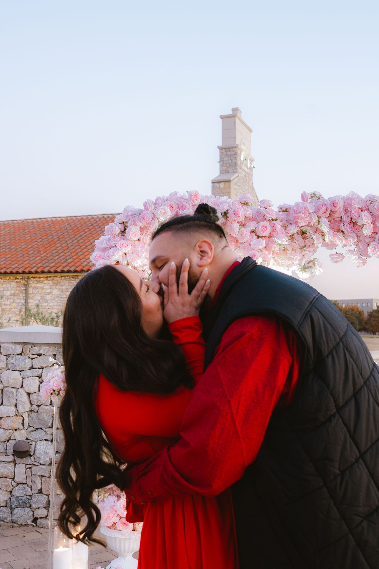 Couple sharing a romantic kiss under a pink floral heart arch during a marriage proposal at sunset.