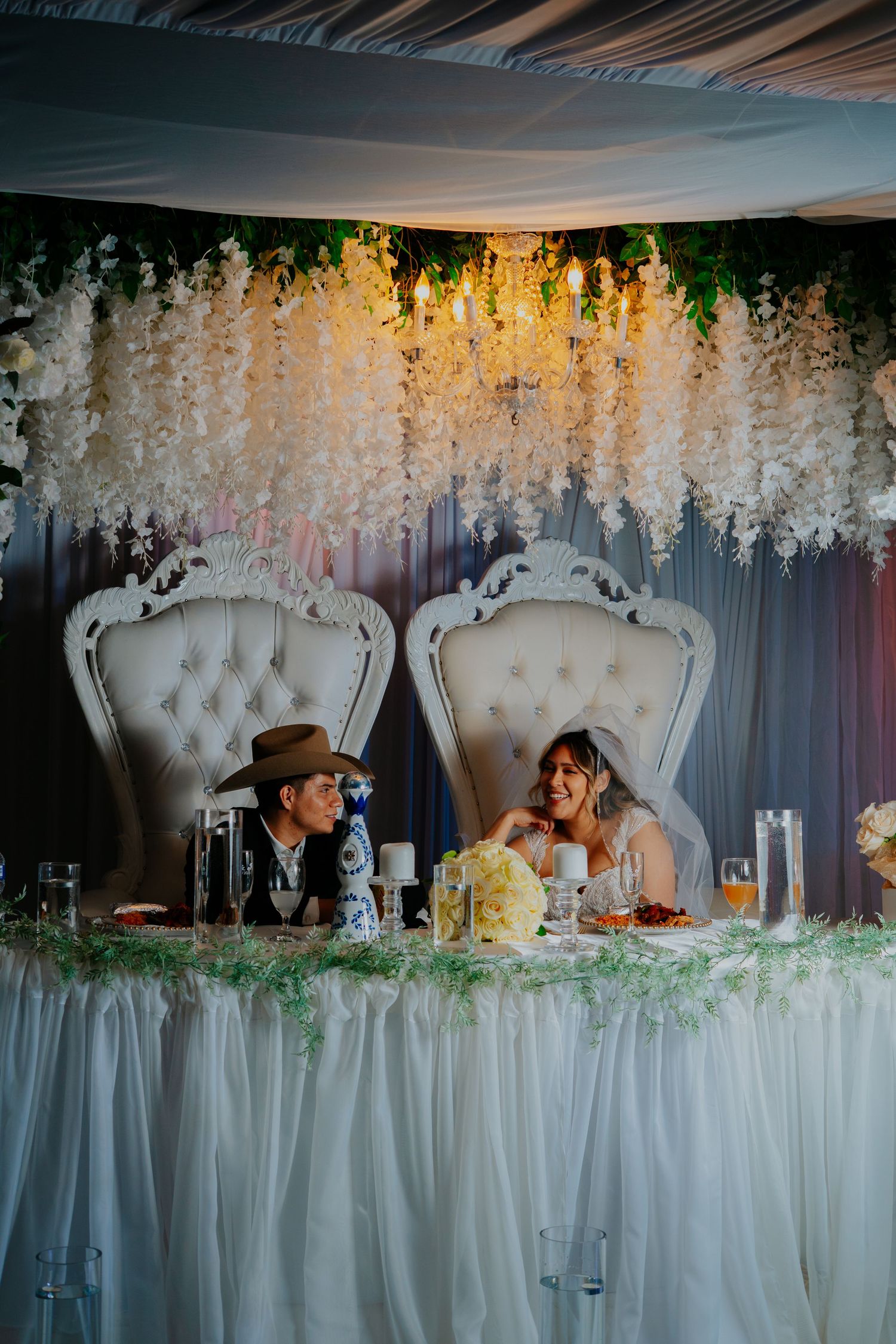 Wedding reception table decorated with white floral arrangements and elegant upholstered chairs under draped fabric ceiling.