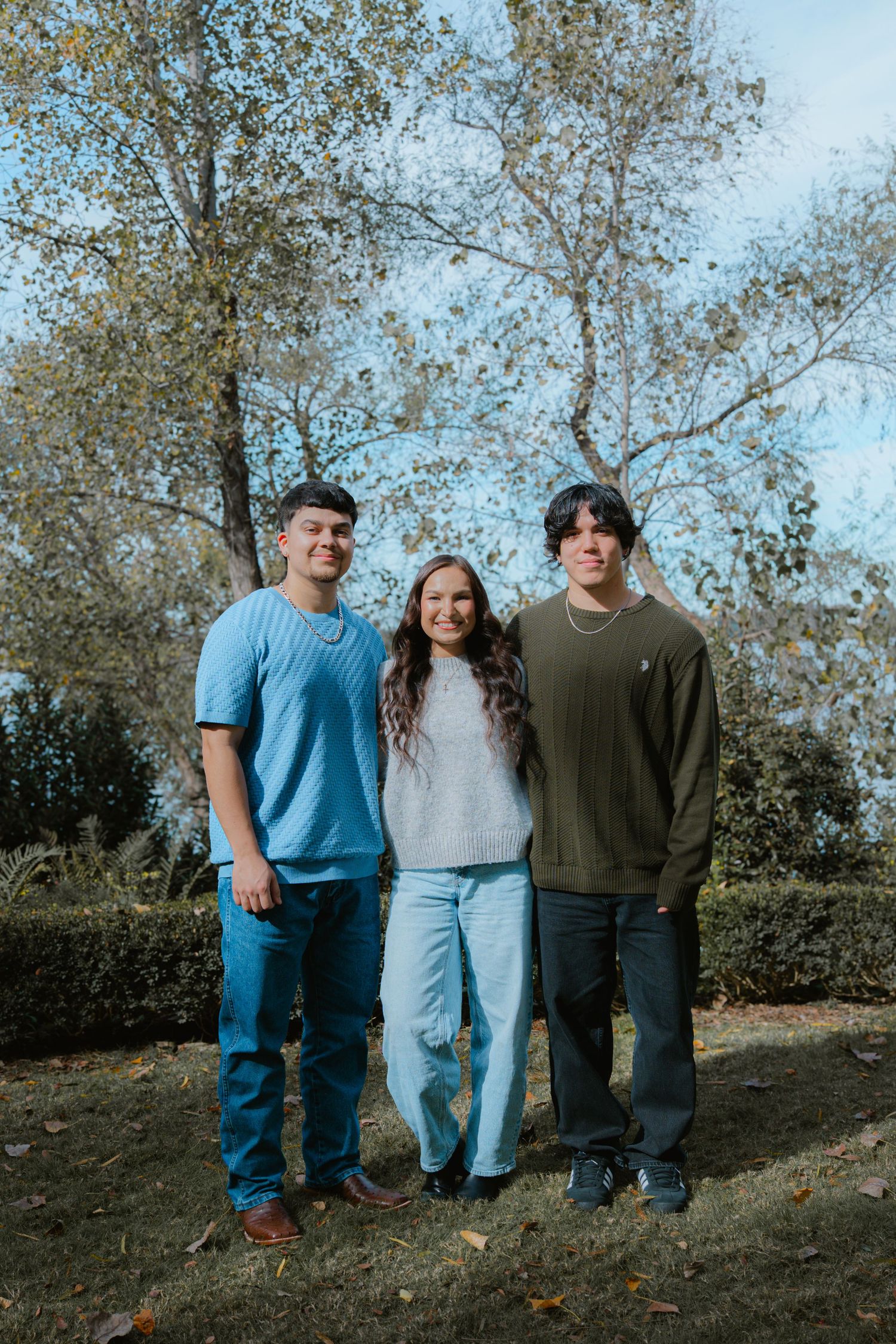 Three people stand together on a grassy hillside with autumn trees in the background.