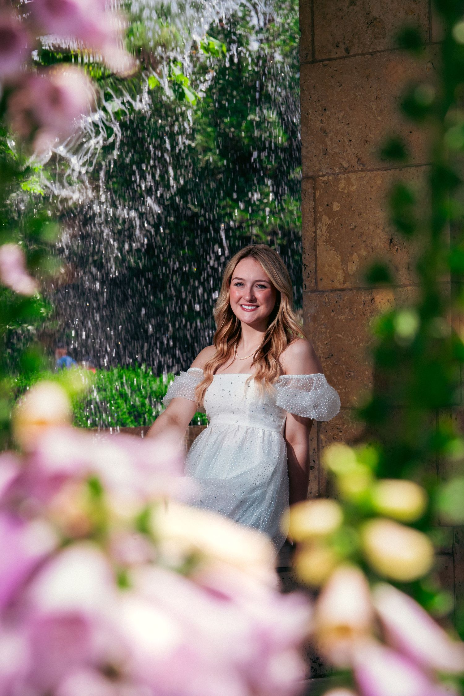Dreamy outdoor portrait in white dress framed by pink flowers and water droplets in a garden setting.