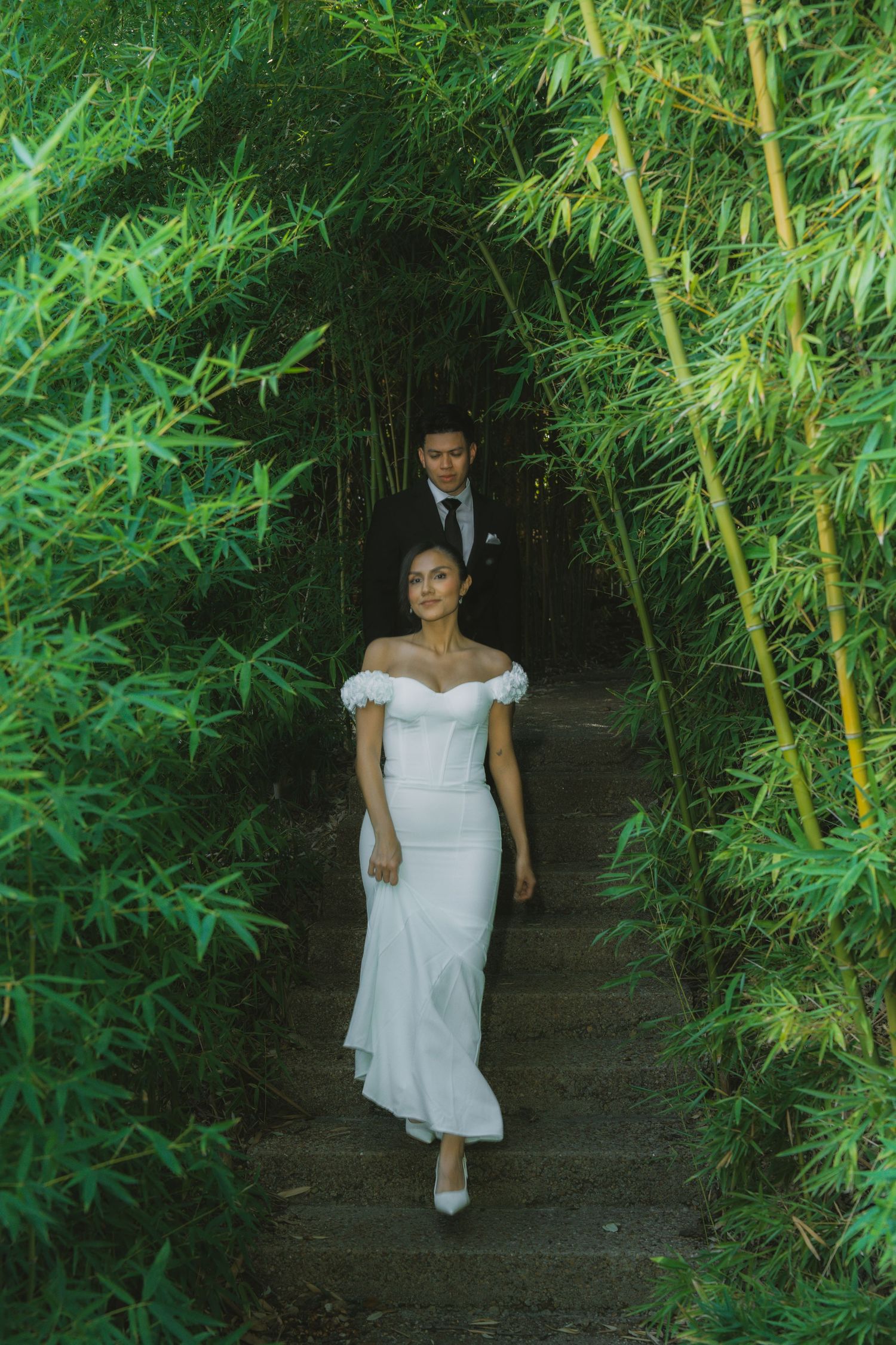 Bride in white off-shoulder gown and groom in black suit walking through lush bamboo garden path.