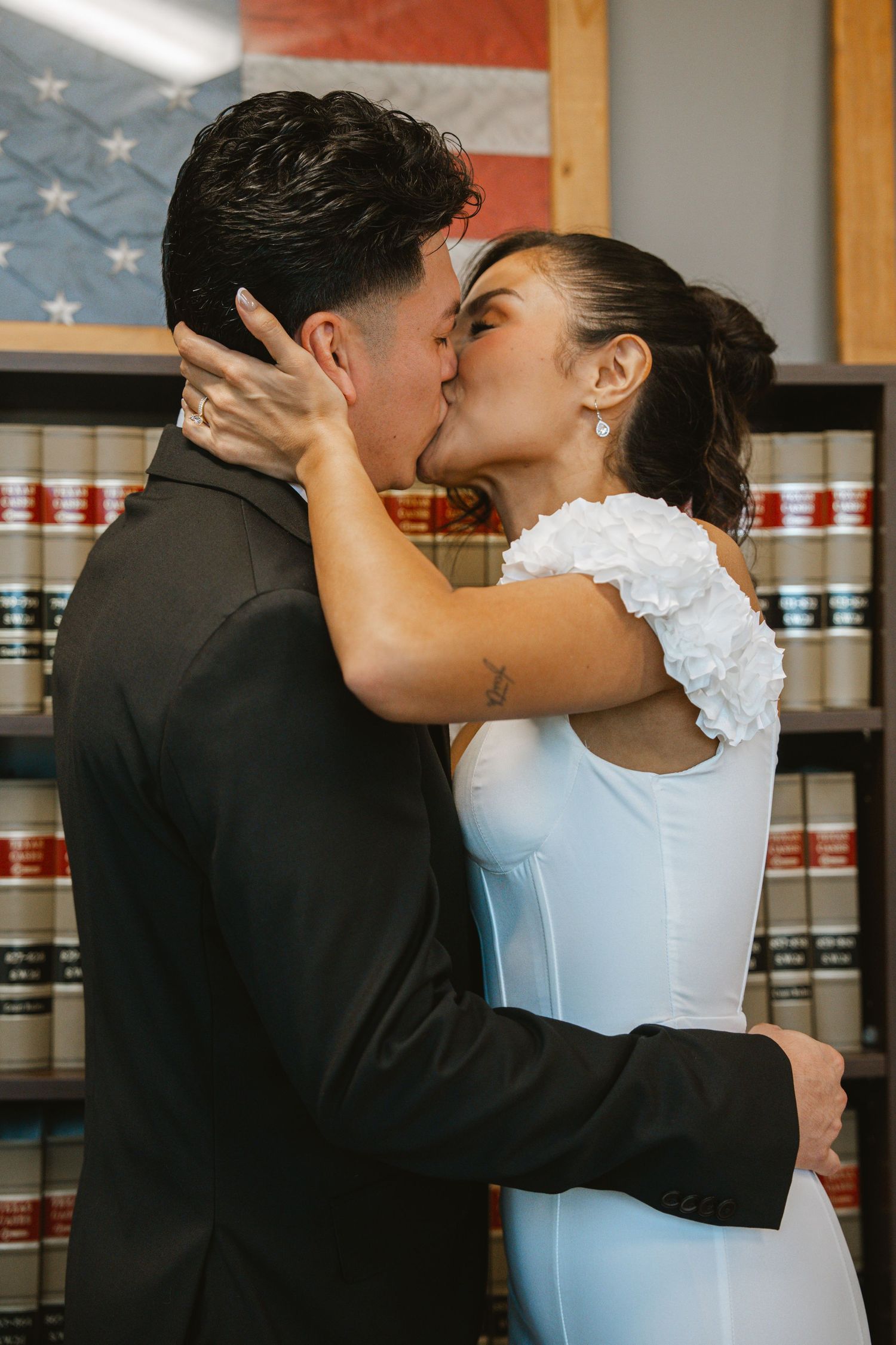 Couple sharing a first kiss in a courthouse wedding, bride in white dress, groom in black suit, American flag background.