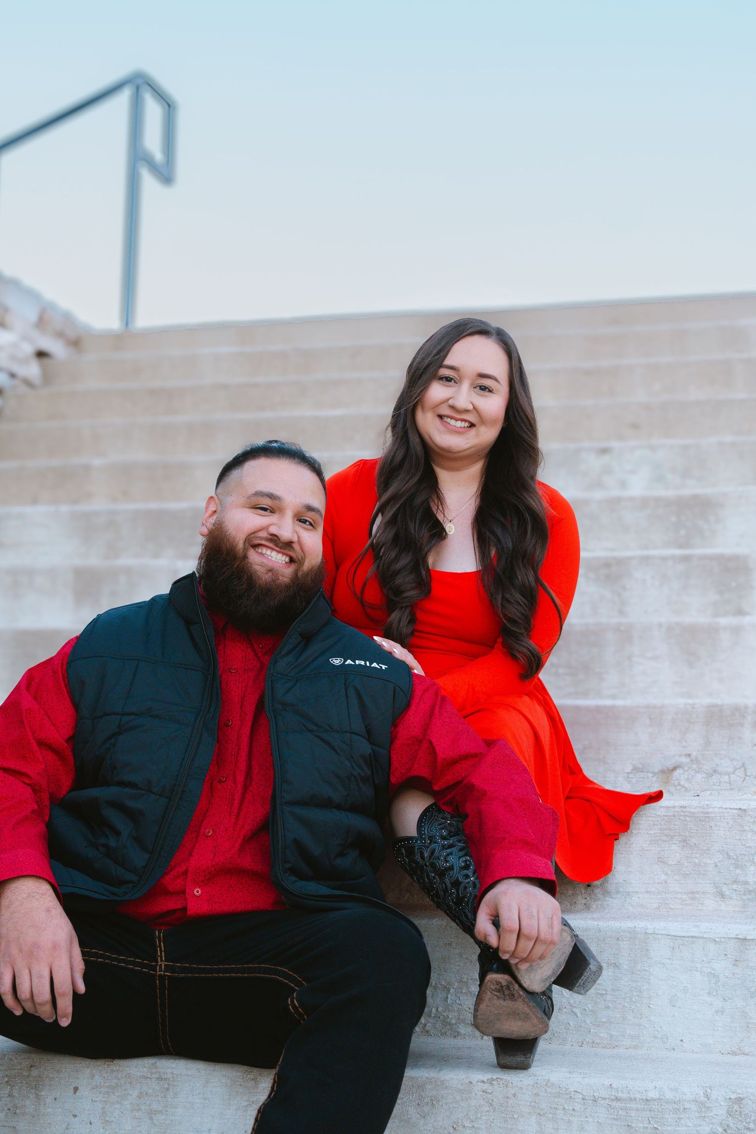Couple in matching red outfits sitting on stone steps, man in black vest smiling at camera, woman in red dress behind him.
