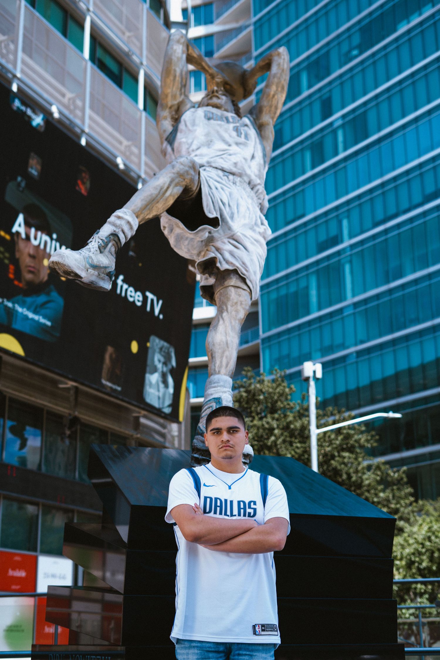 Young man in Dallas Mavericks jersey stands arms crossed before a large basketball player statue outdoors.