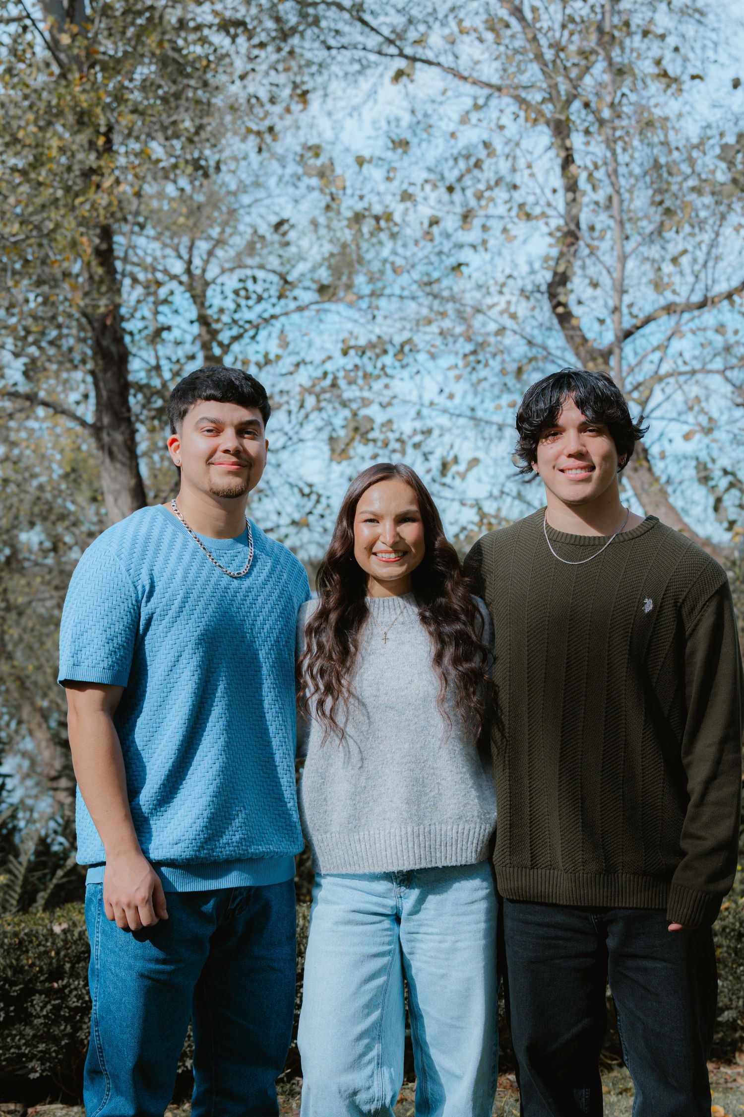 A group of three people wearing casual sweaters and t-shirts pose together outdoors against a backdrop of autumn trees.