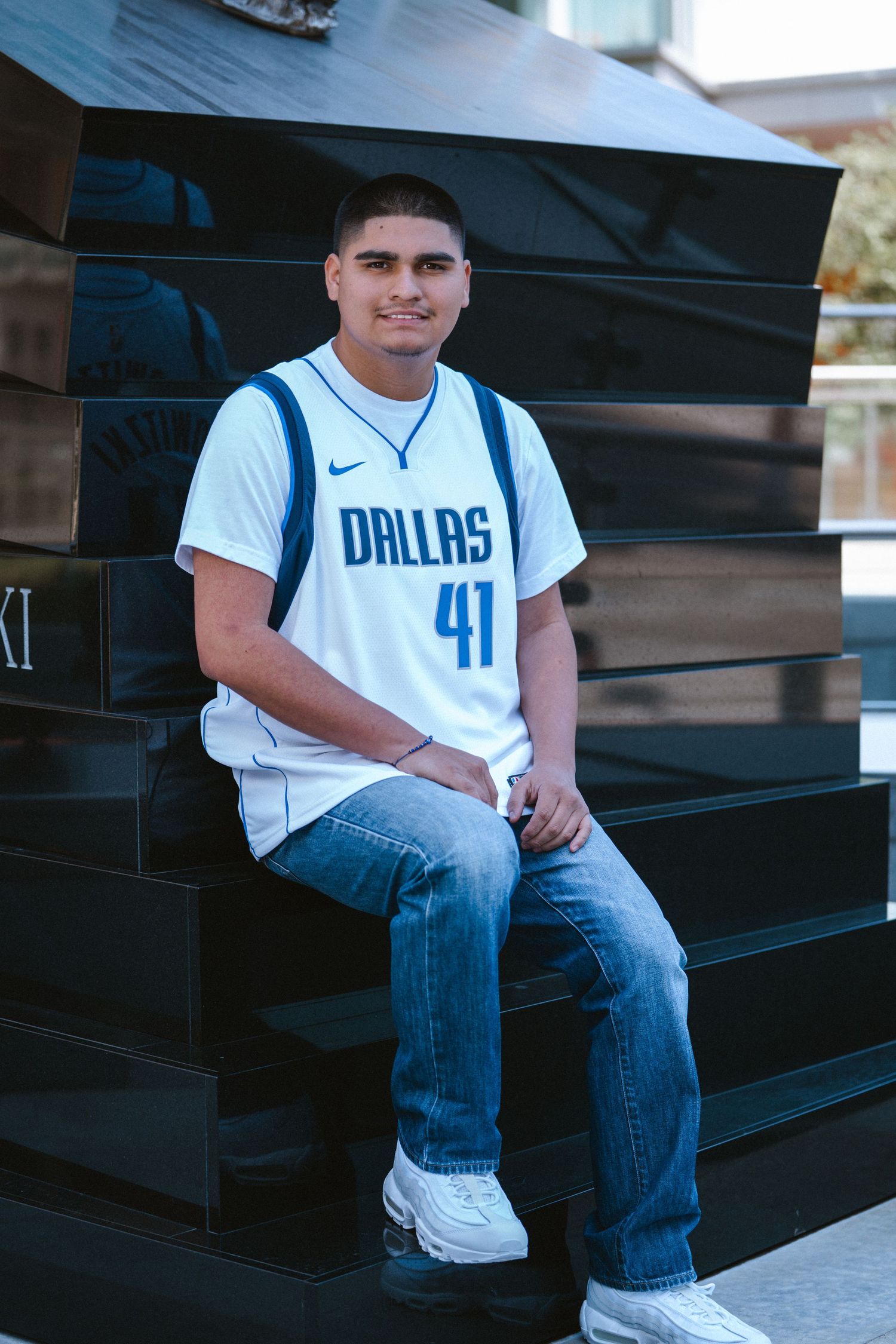 Young man in Dallas Mavericks #41 jersey and jeans sitting on dark steps outdoors smiling.