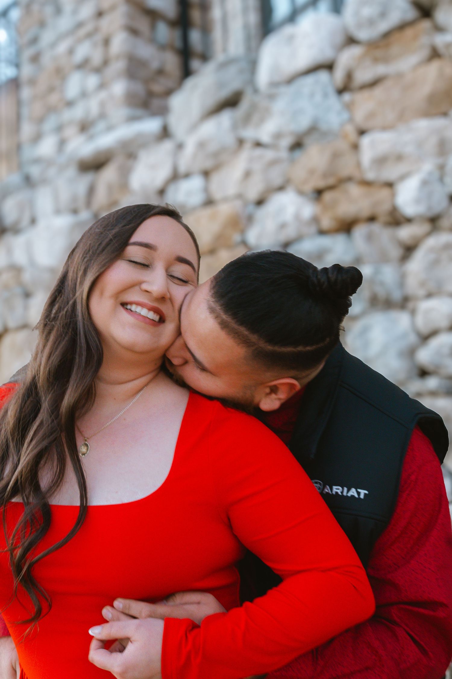 Couple in matching red outfits sharing a romantic kiss against a rustic stone wall backdrop.