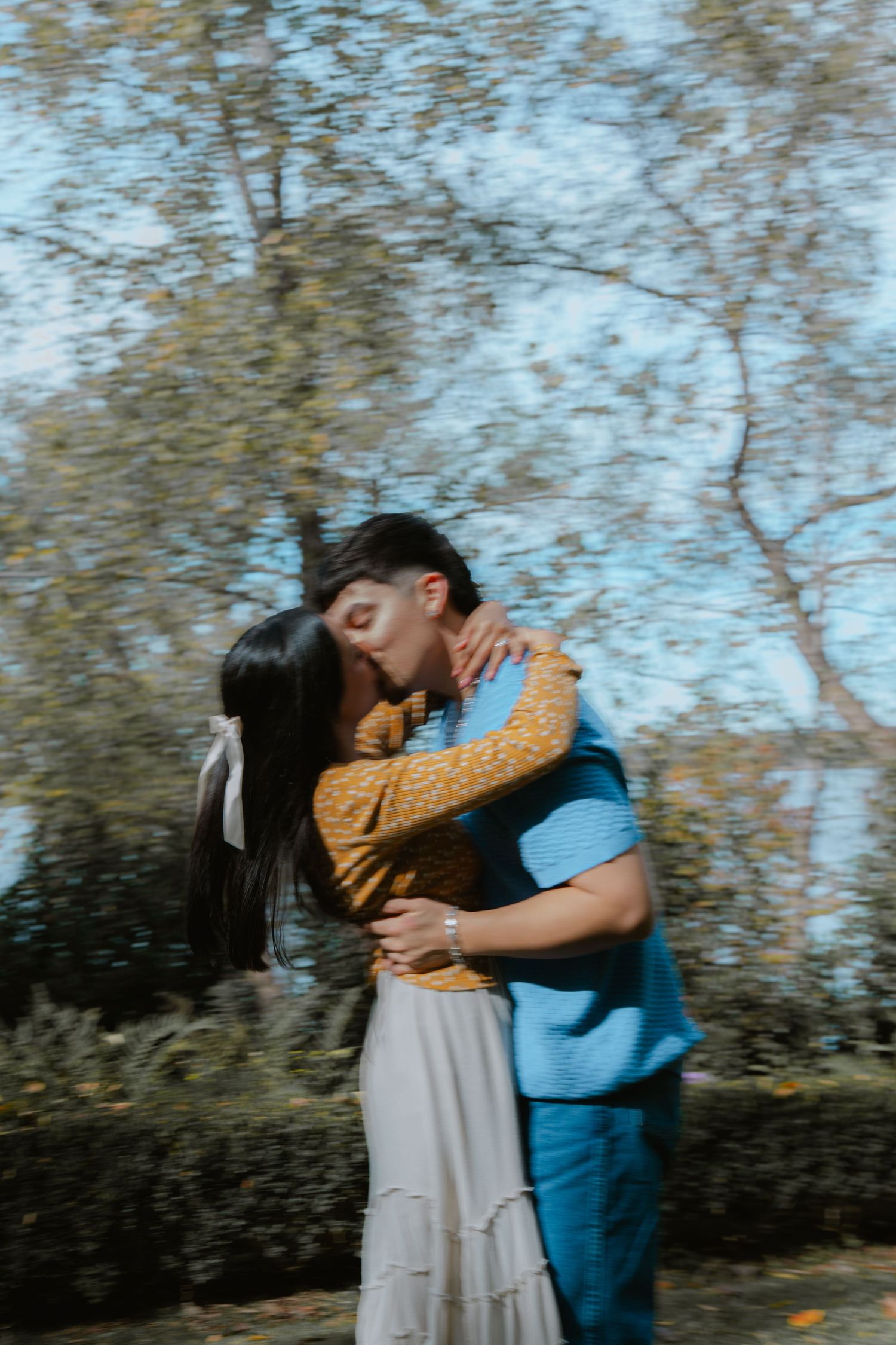 A couple in casual attire sharing an intimate embrace outdoors against a backdrop of trees and sky.