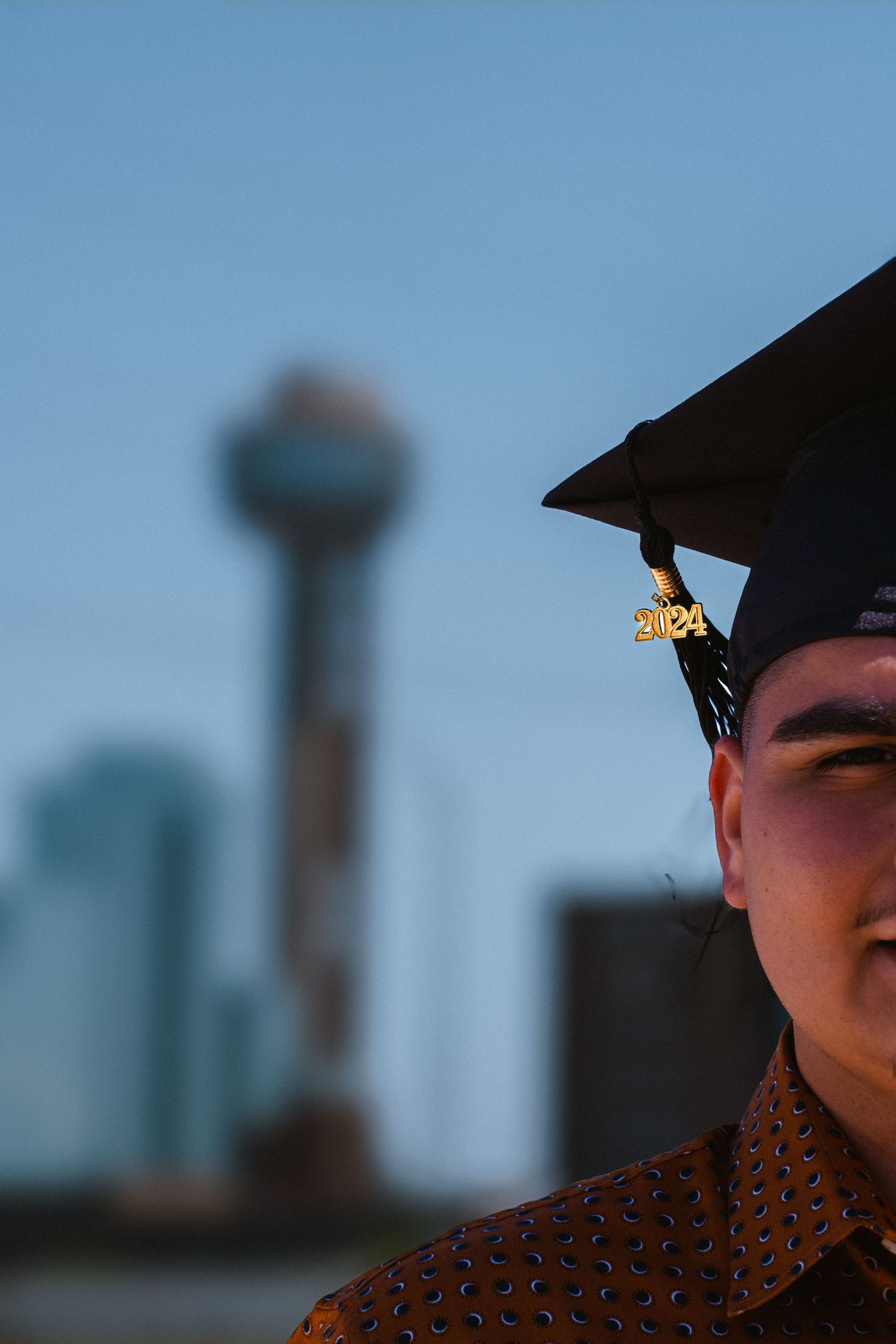 Graduate in 2024 mortarboard cap poses in front of blurred Reunion Tower in Dallas skyline at dusk.