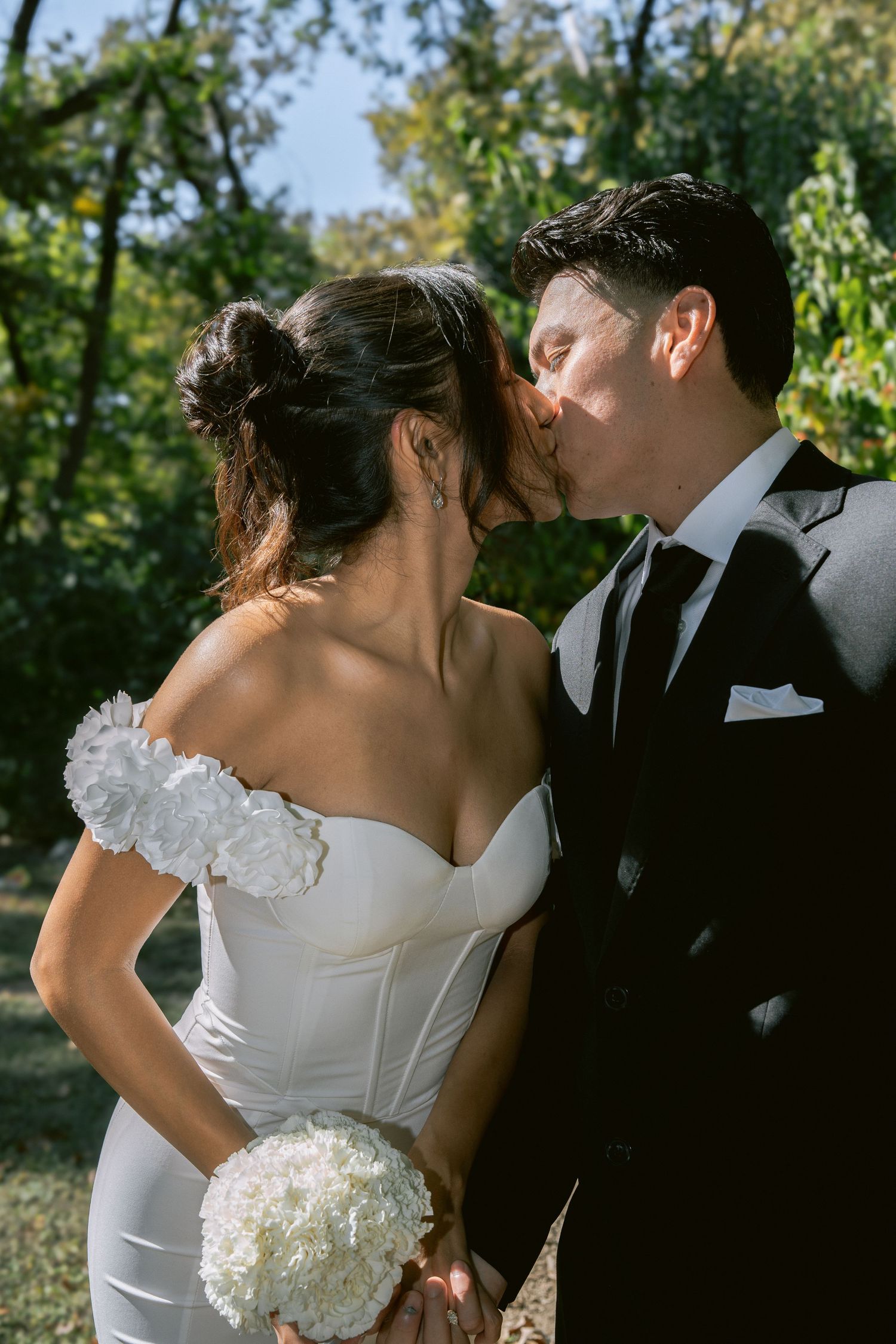 A bride and groom share a romantic kiss outdoors, she holds white hydrangeas wearing an off-shoulder gown, he wears a black suit.