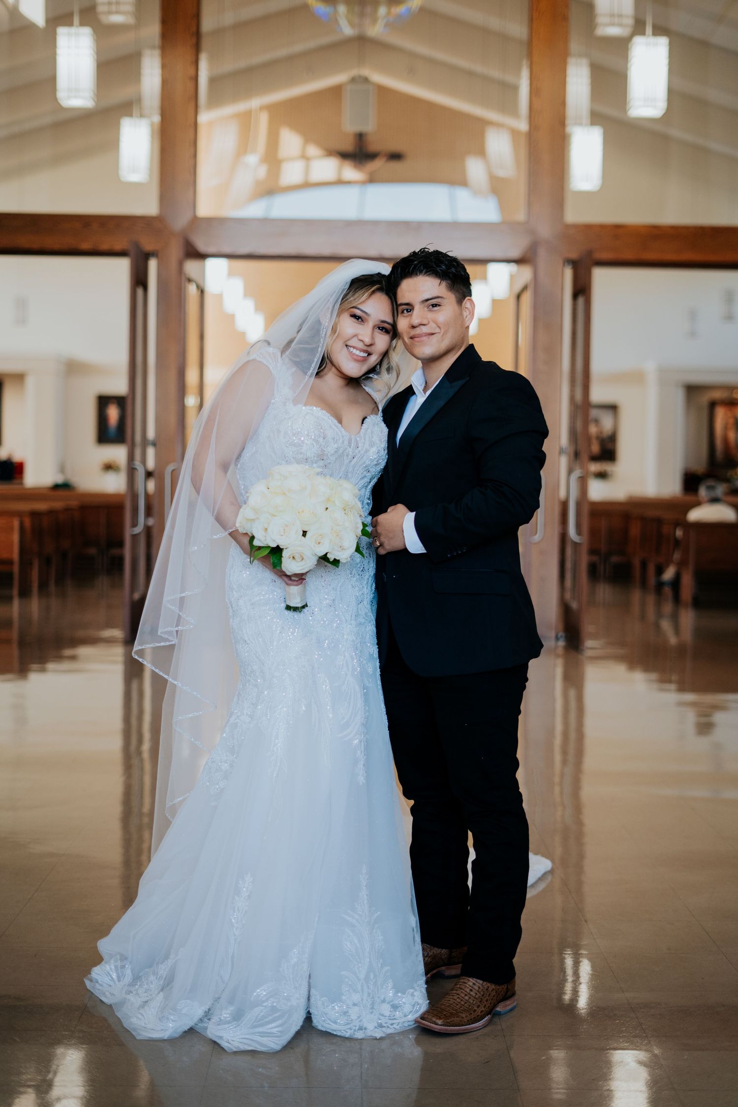 A couple poses together in a church hall wearing formal wedding attire with white decor and pendant lights.