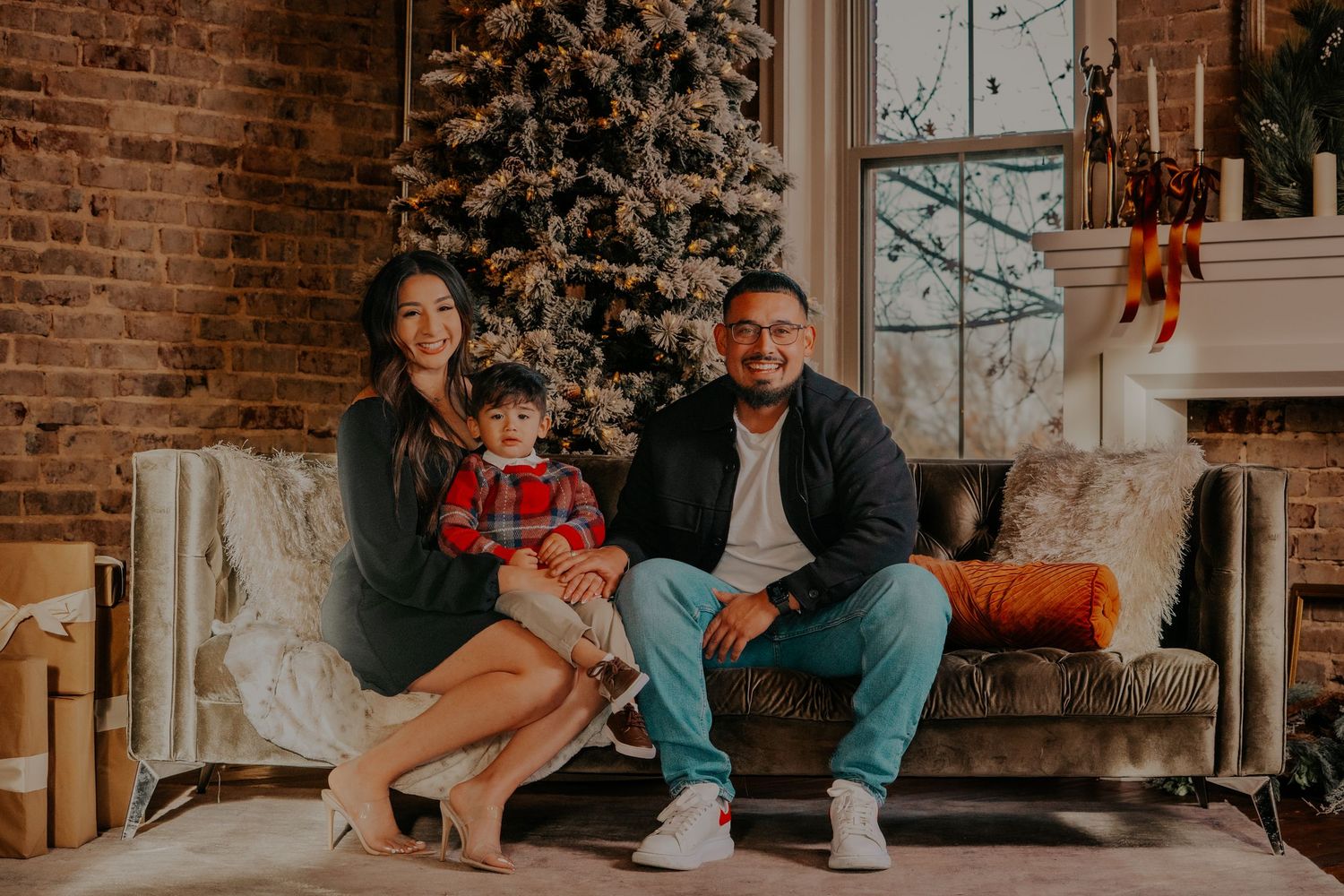 Family sits together on brown leather couch in rustic brick loft with large windows and holiday decorations.