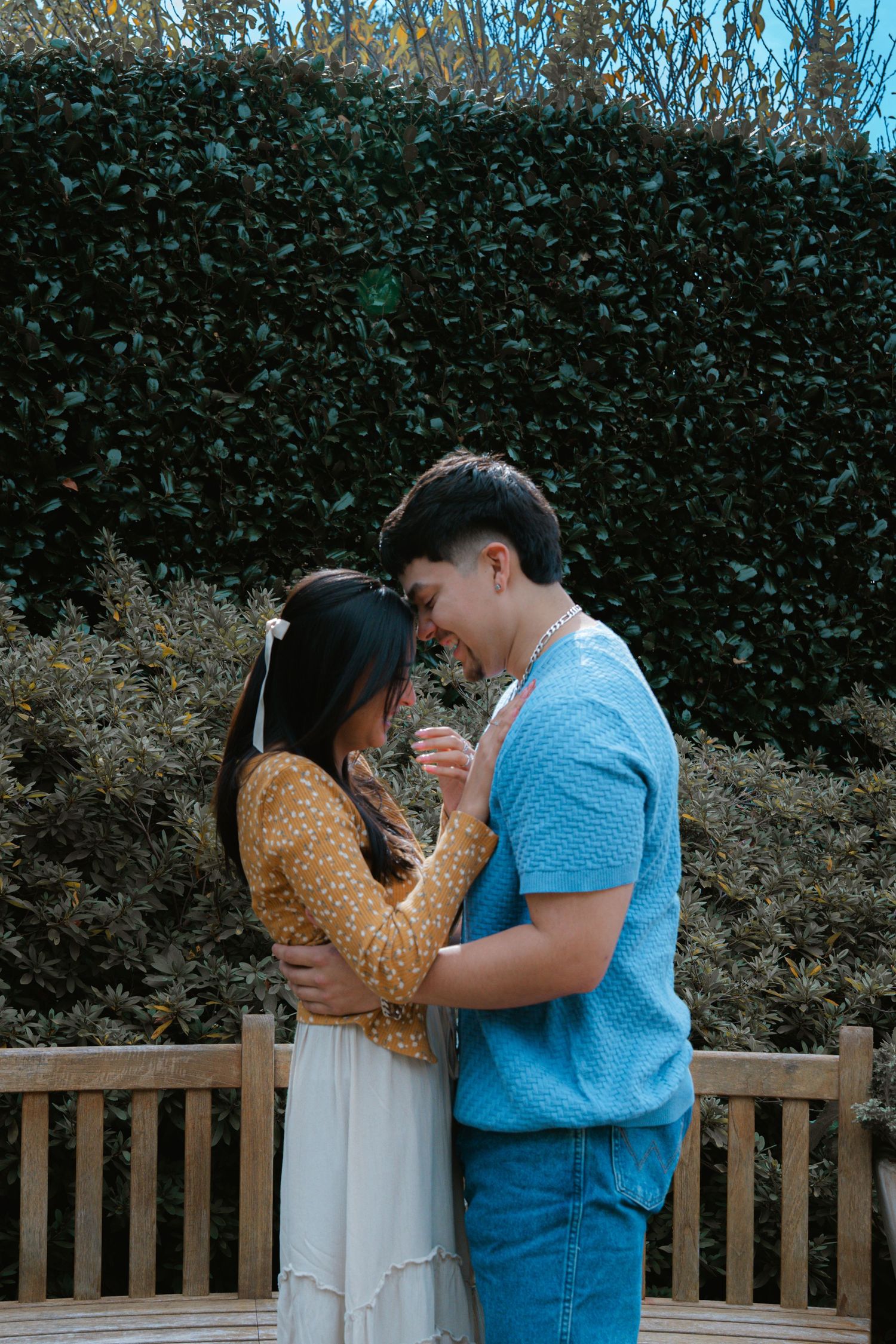 A couple embraces in a romantic moment outdoors in front of a green hedge.