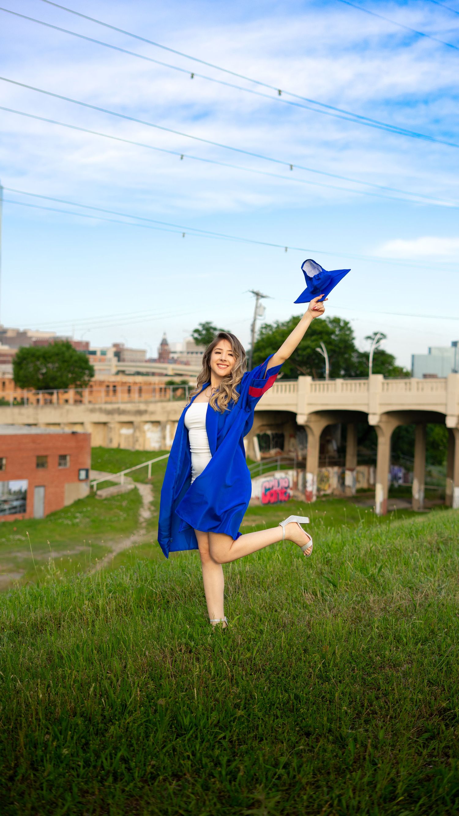 A dancer in a flowing blue dress performs graceful movements on a grassy hillside near brick buildings and a bridge.