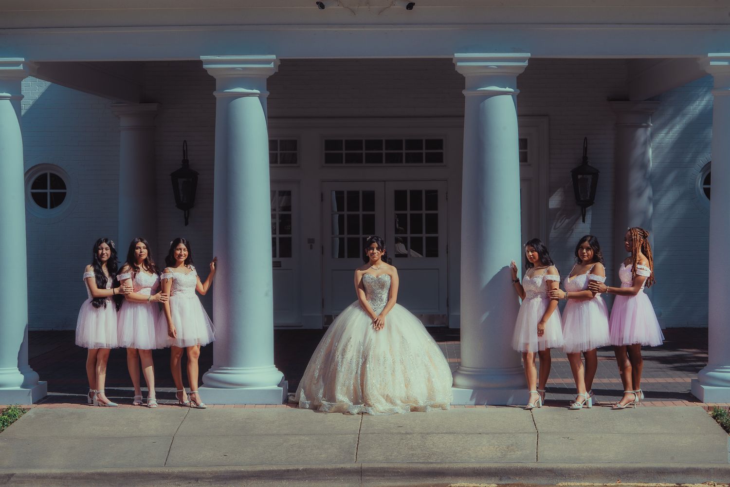 Bride in white ball gown poses with bridesmaids in pink dresses before elegant white columned venue.