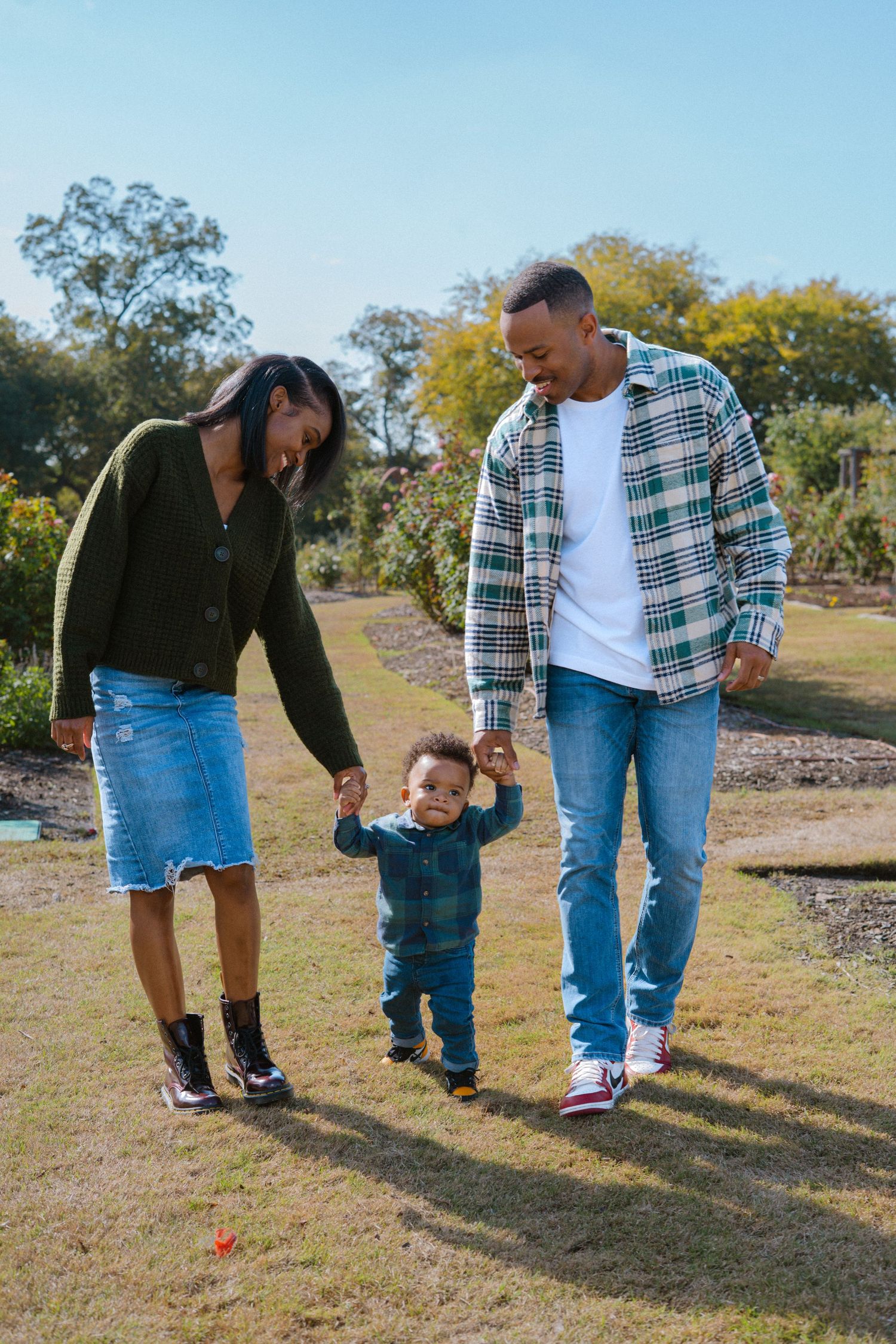 A young Black family walks hand-in-hand with their toddler in a sunny park, wearing casual denim and plaid outfits.