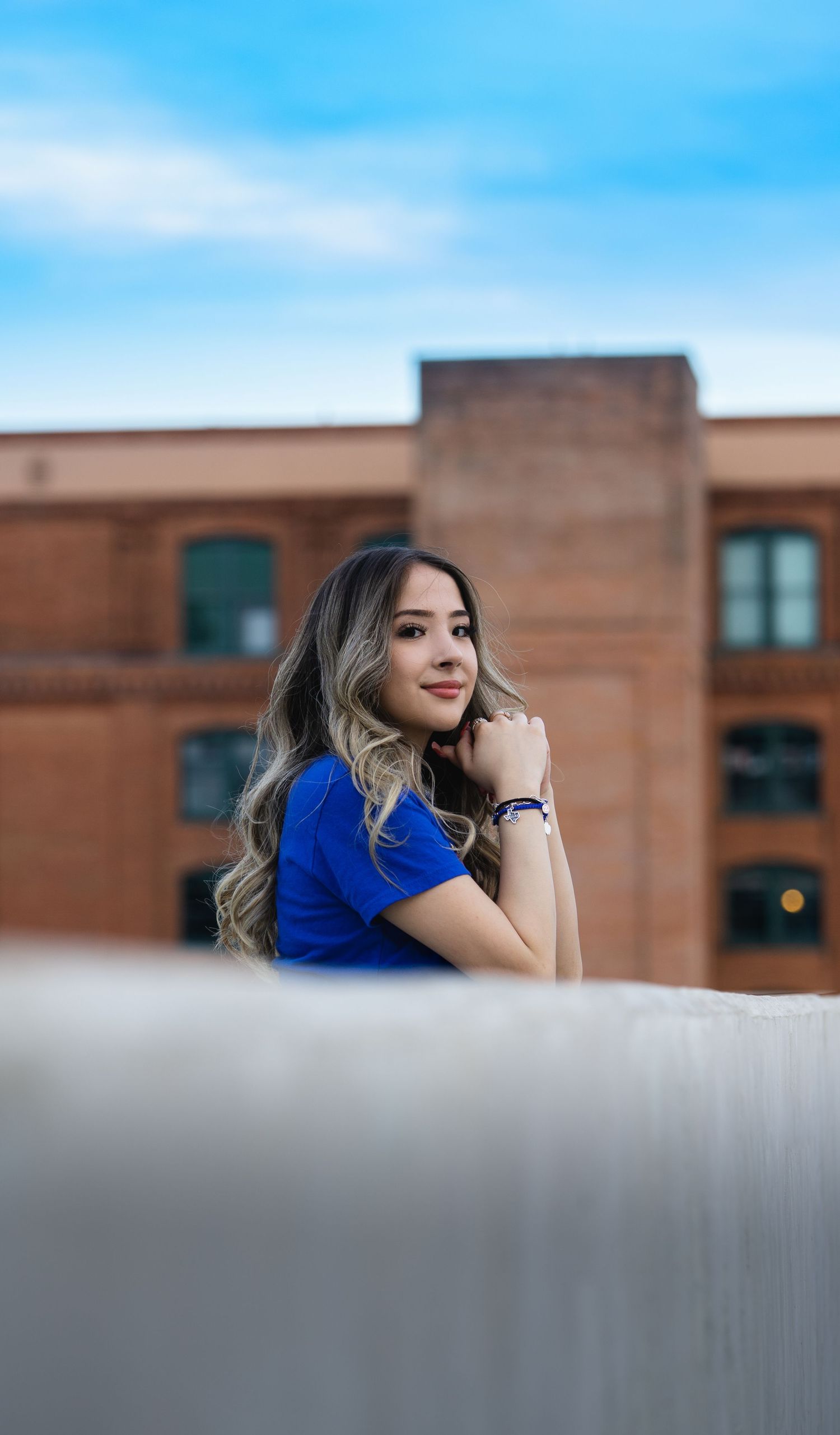 Person in bright blue top poses against blurred background with brick building and blue sky.
