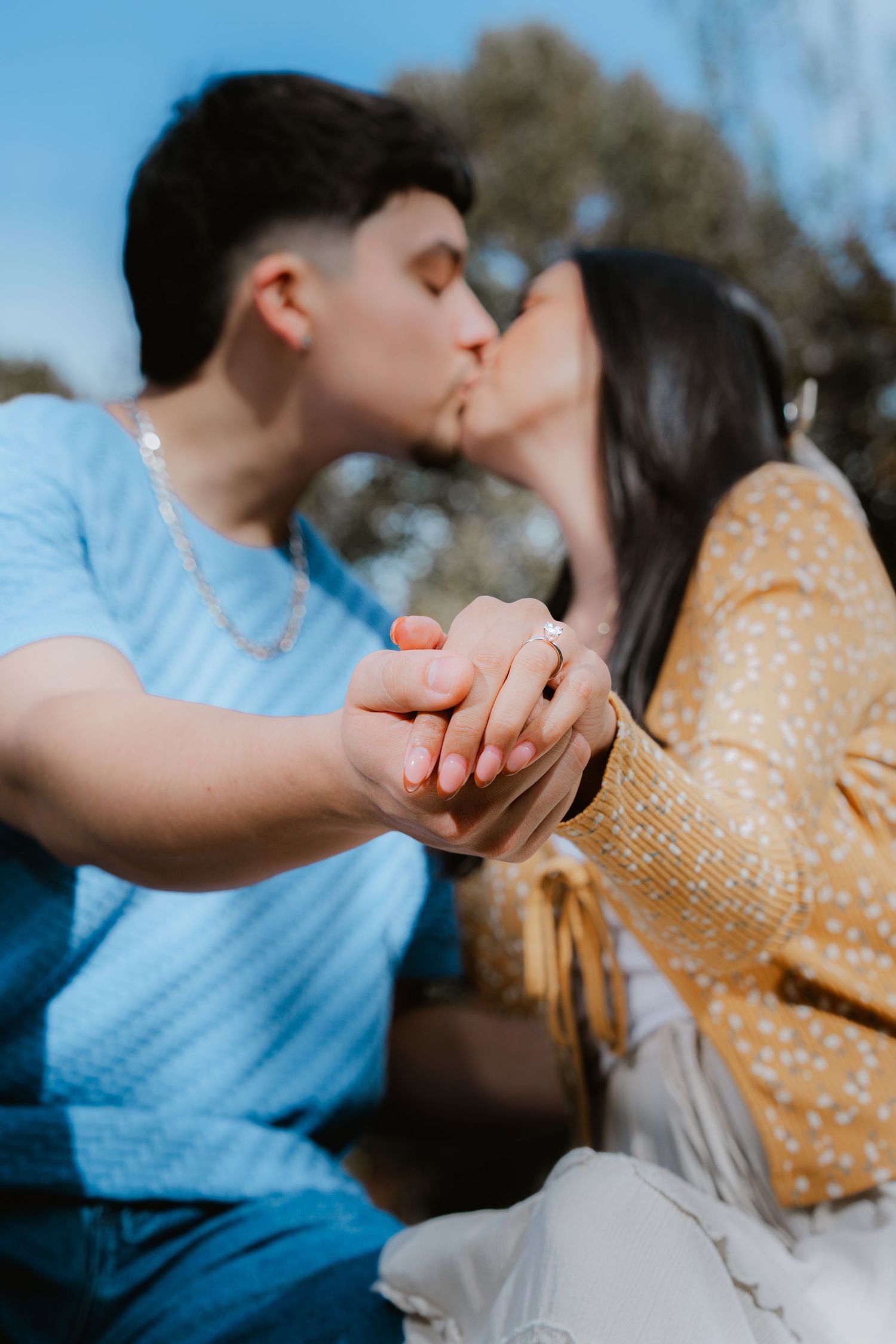 A romantic couple holds hands while sharing a tender kiss outdoors on a sunny day.