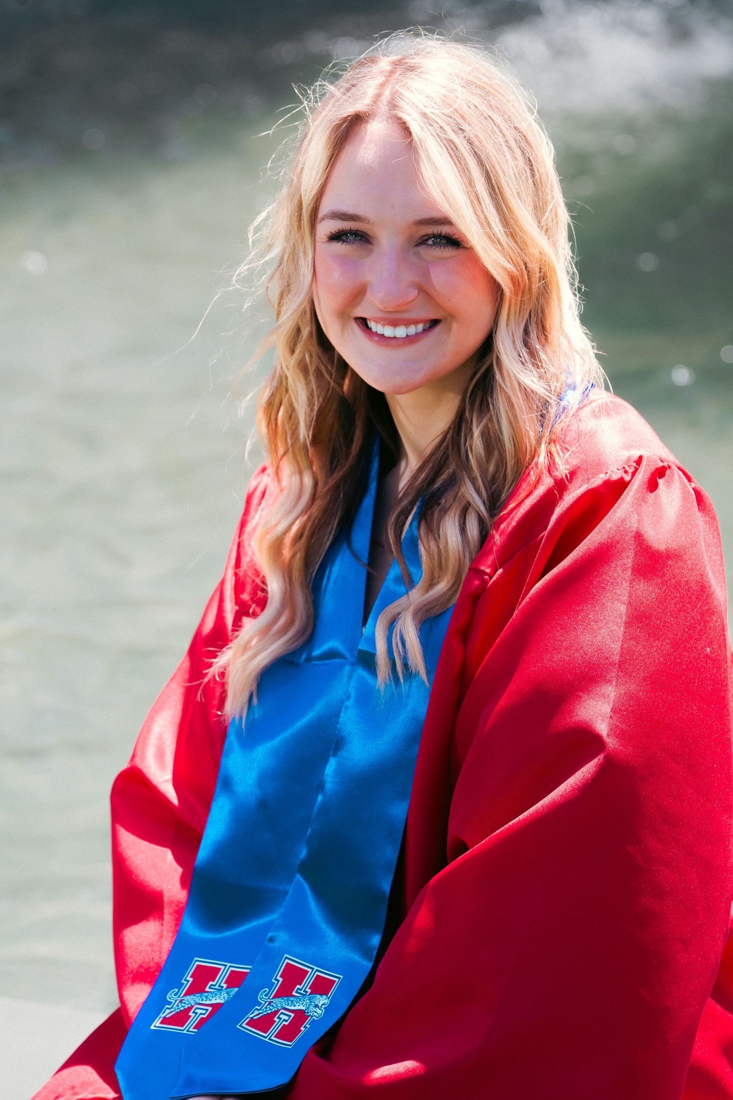 A person wearing a bright red coat and blue top poses outdoors against a soft, blurred natural background.
