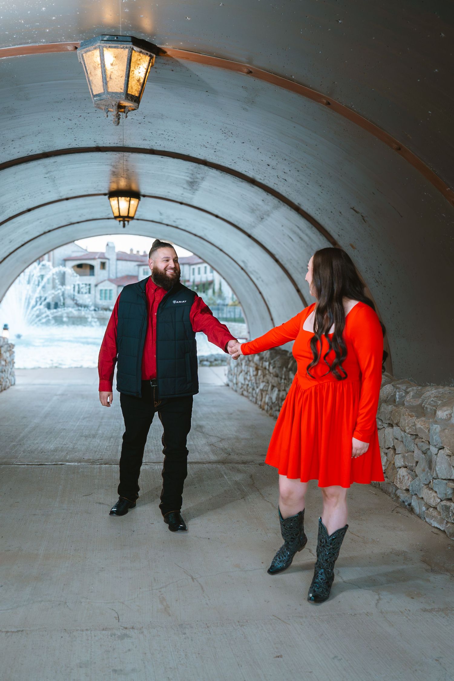 Couple holding hands in an arched tunnel, woman in red dress and boots, man in black vest and red shirt.