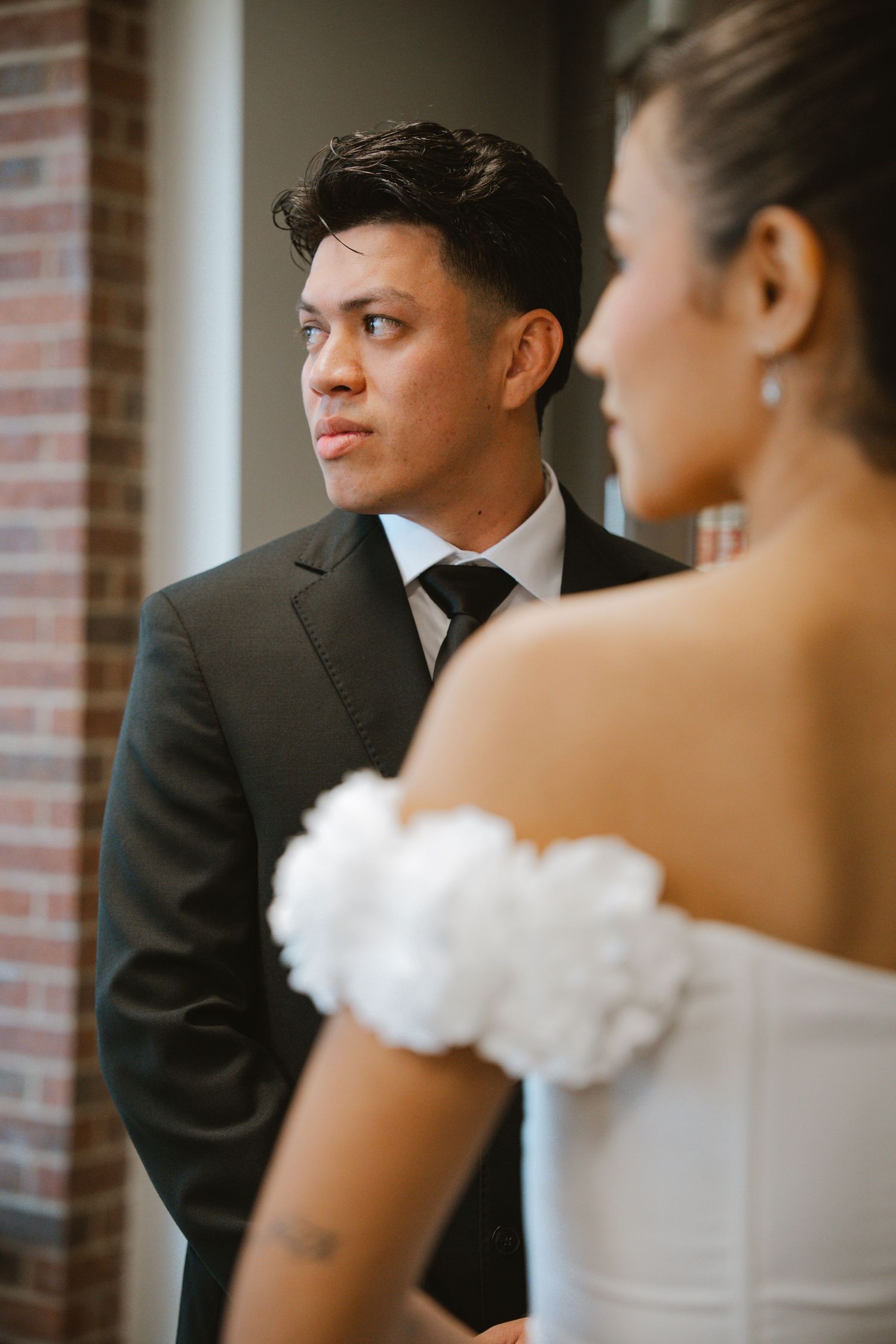 Groom in black suit and tie gazes thoughtfully as bride in white off-shoulder dress with floral detail stands in foreground.