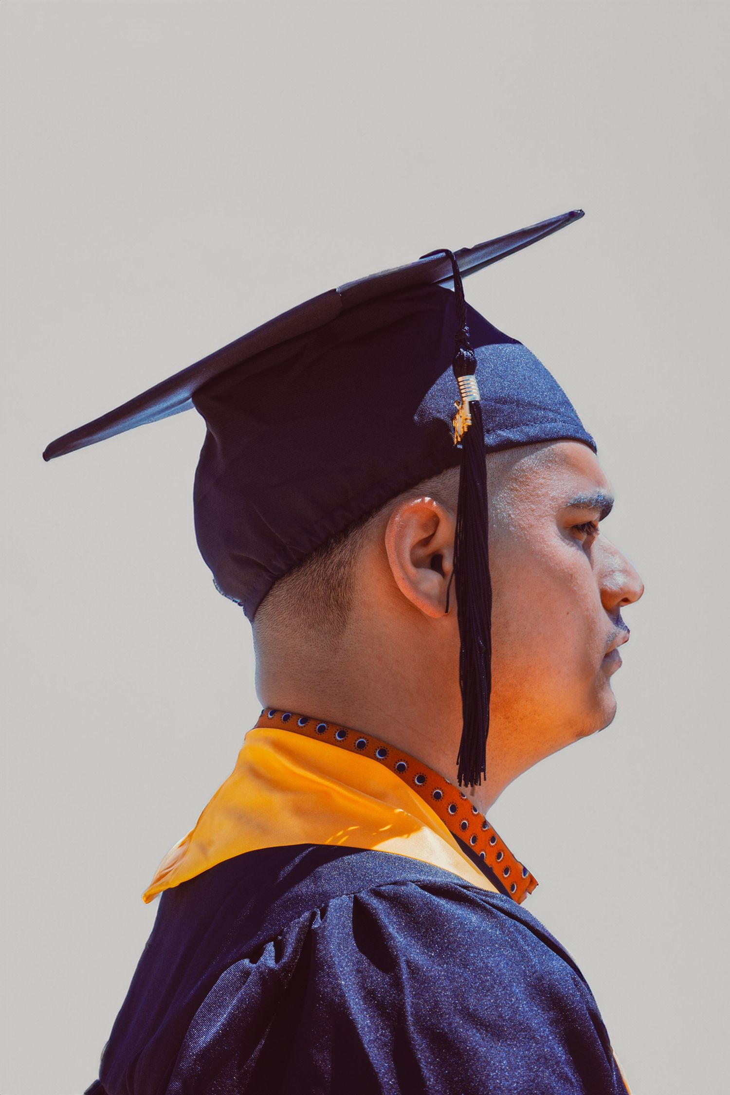 Young man in black graduation cap and gown with gold collar, side profile view against gray background.