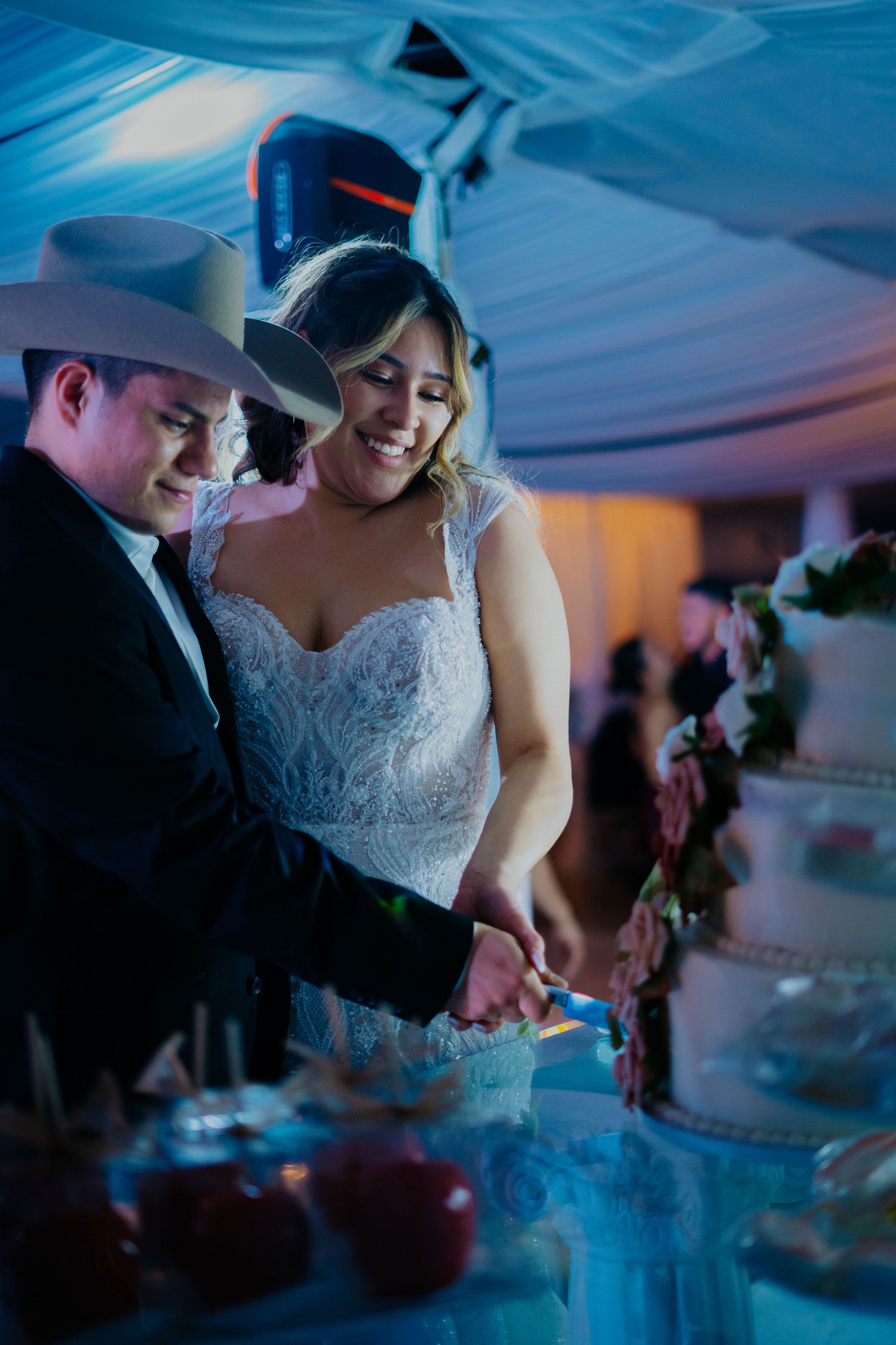 A couple in wedding attire cut their wedding cake together under blue lighting at an evening reception.