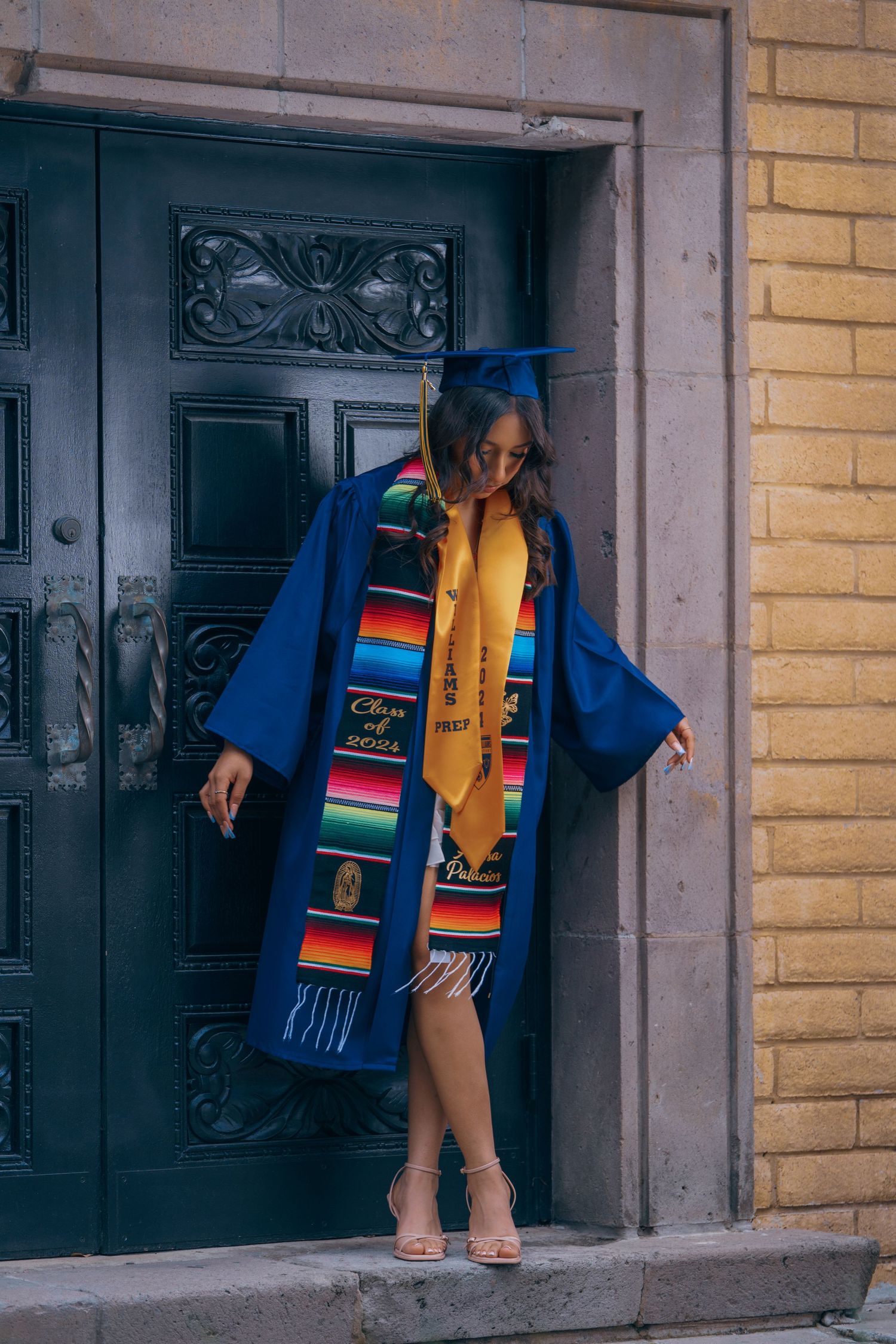 Graduate in blue graduation attire and multicolored stole poses near ornate black door and yellow wall.