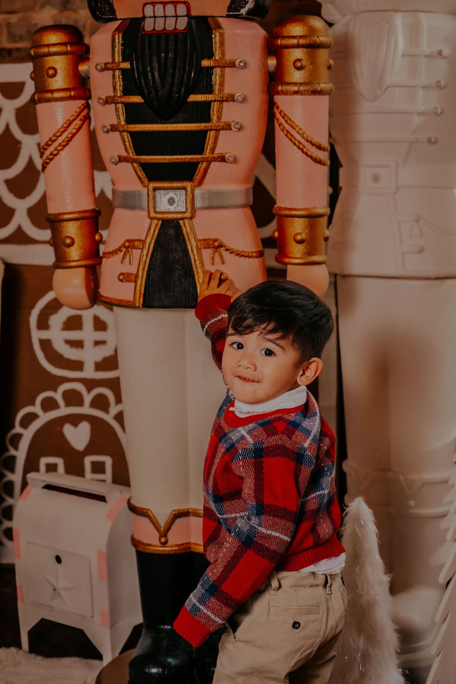 Red plaid and beige holiday outfit against ornate nutcracker decorations and brick wall backdrop.
