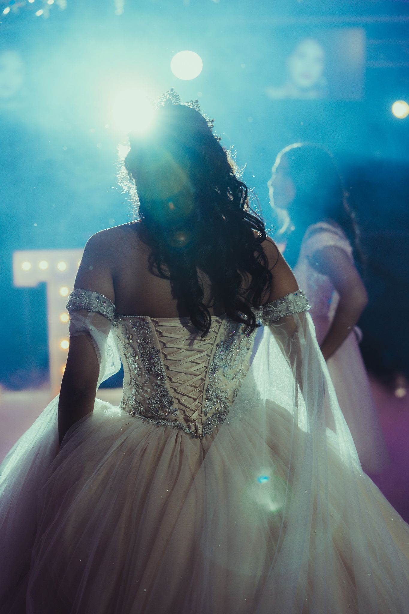 A quinceañera girl in a beaded off-shoulder ball gown with corset back, wearing a tiara, dancing under blue stage lights.