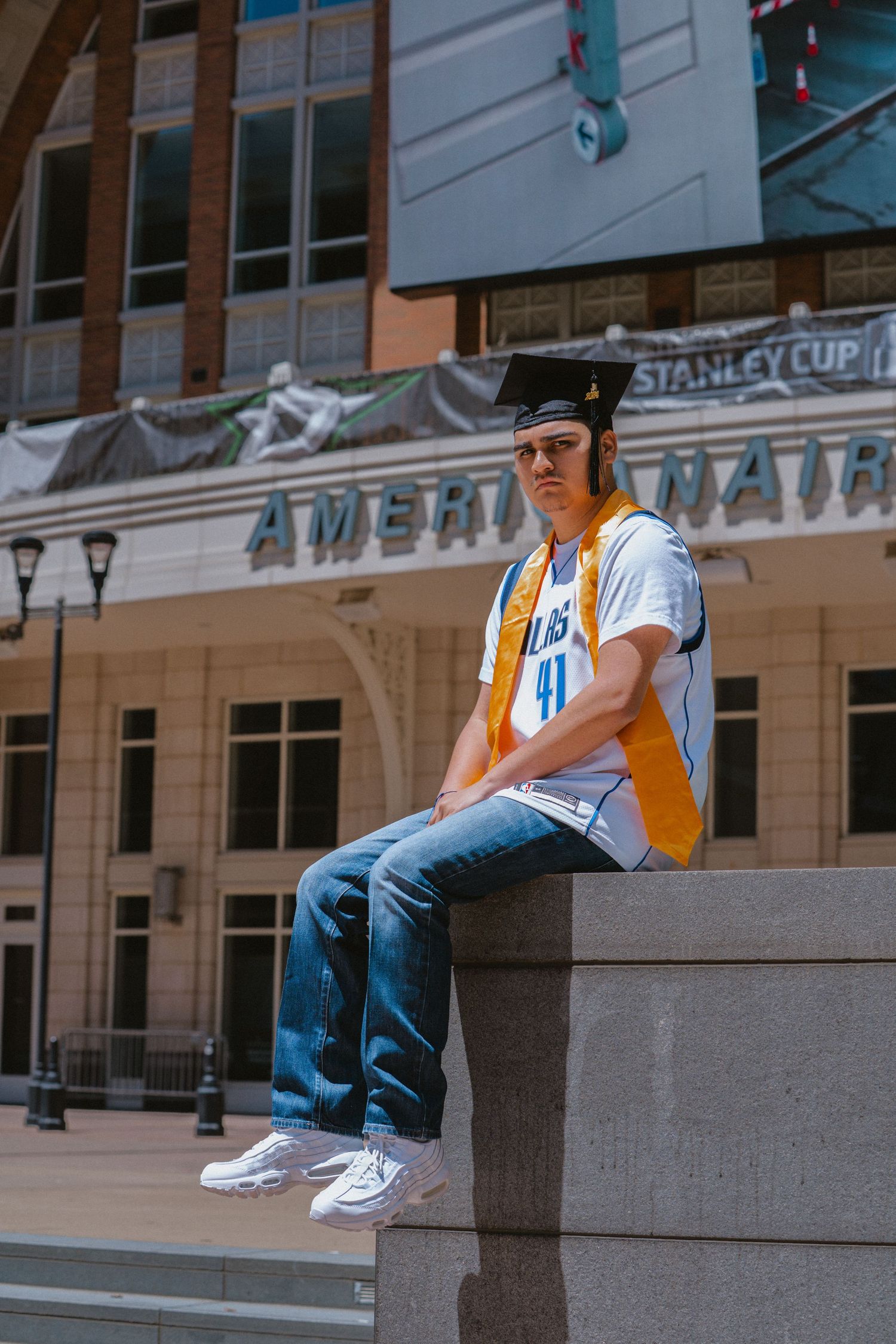 Young graduate wearing cap and gown sash over Mavericks jersey poses outside American Airlines Center.